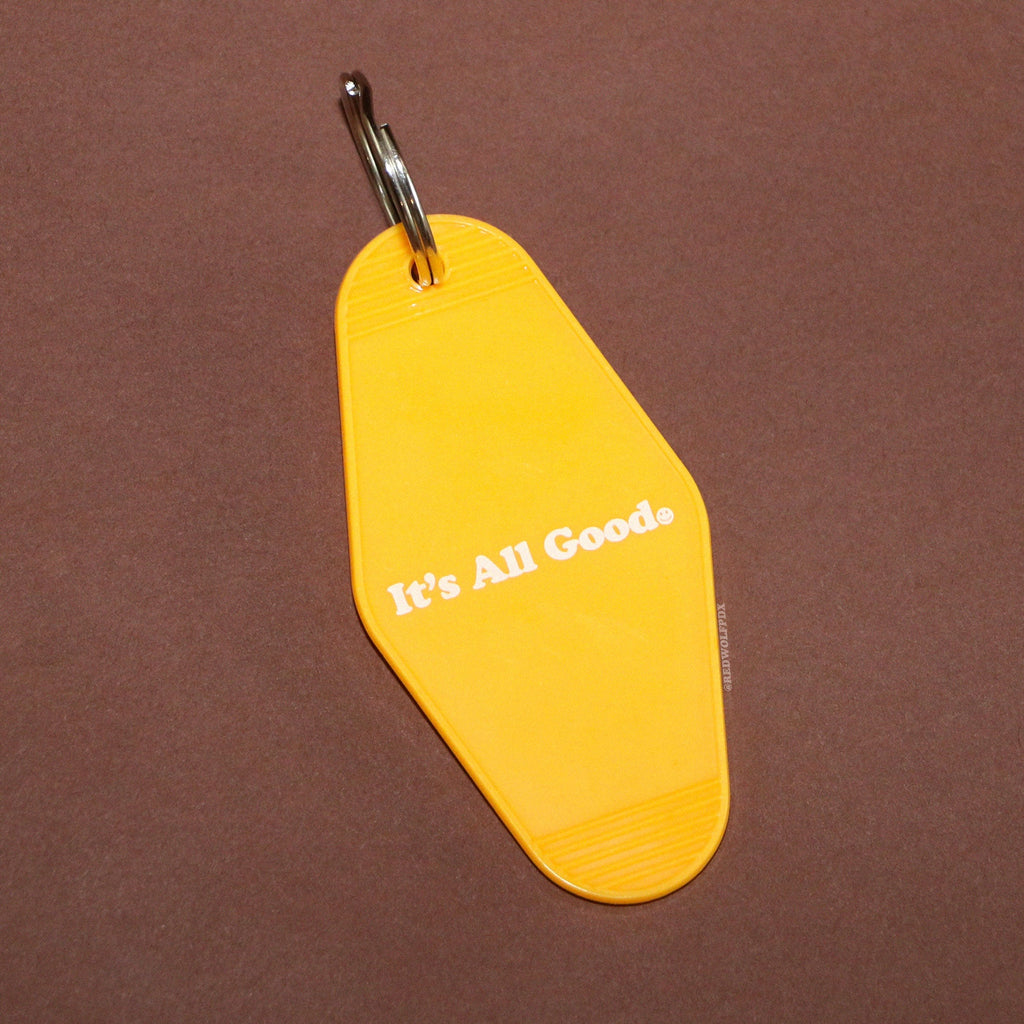 yellow retro motel style keychain with text 'It's All Good' with smiley face in white ink printed across it on a brown background