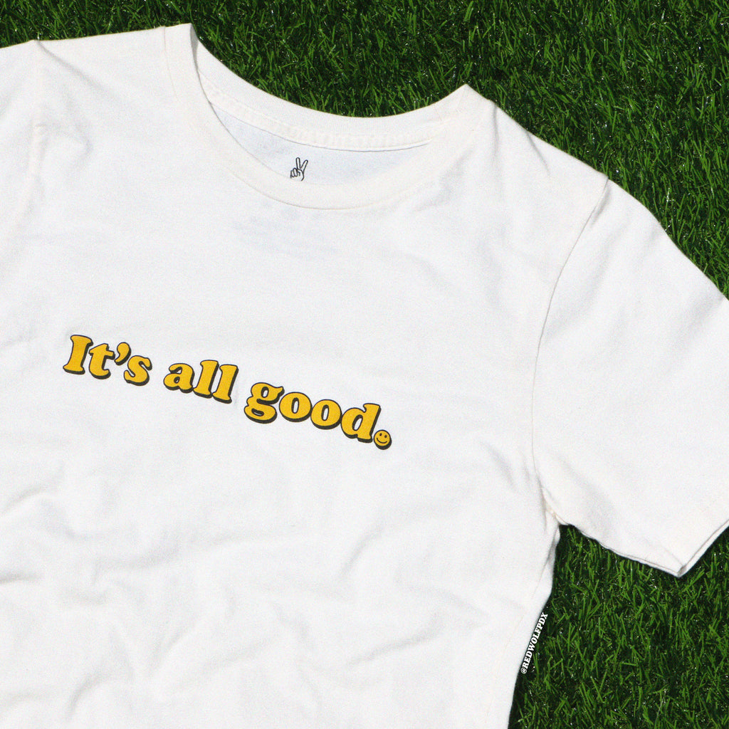 beige graphic tee with text 'It's all good' with smiley face illustration on  green grass background