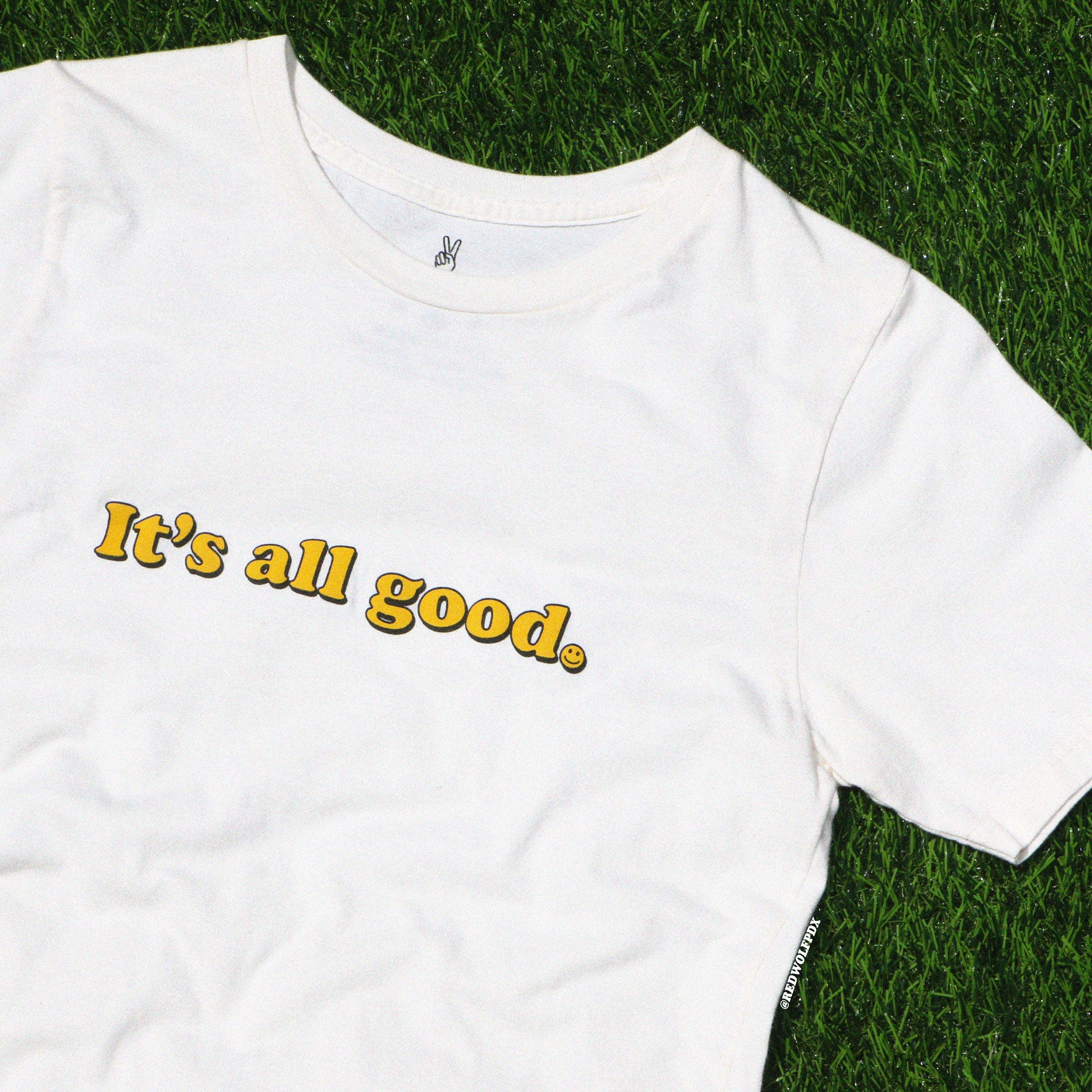 beige graphic tee with text 'It's all good' with smiley face illustration on  green grass background