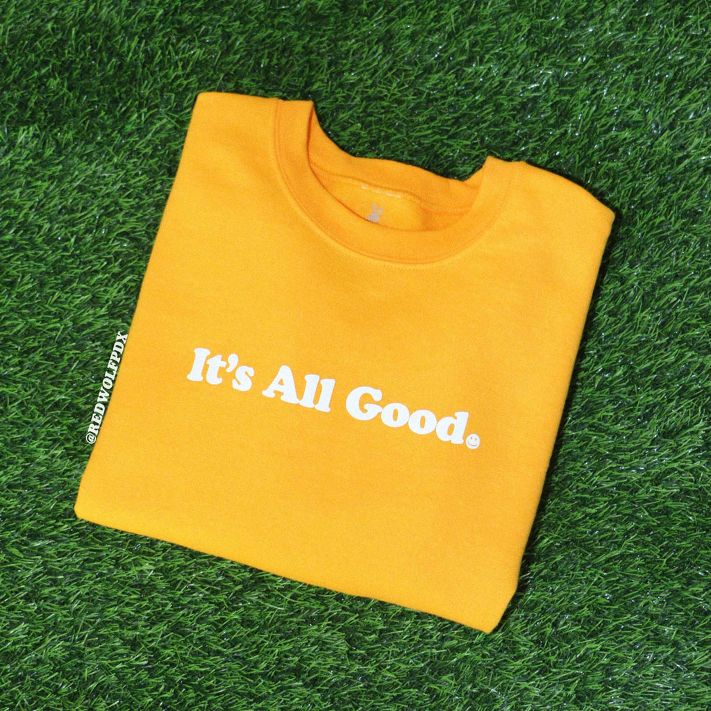 Folded yellow crewneck sweatshirt with text 'It's All Good' with smiley face graphic screen printed in white ink across chest on green grass