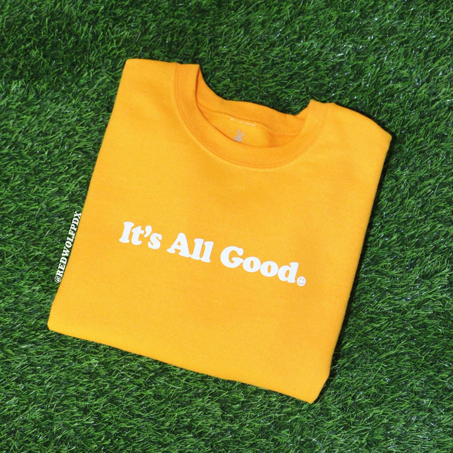 Folded yellow crewneck sweatshirt with text 'It's All Good' with smiley face graphic screen printed in white ink across chest on green grass
