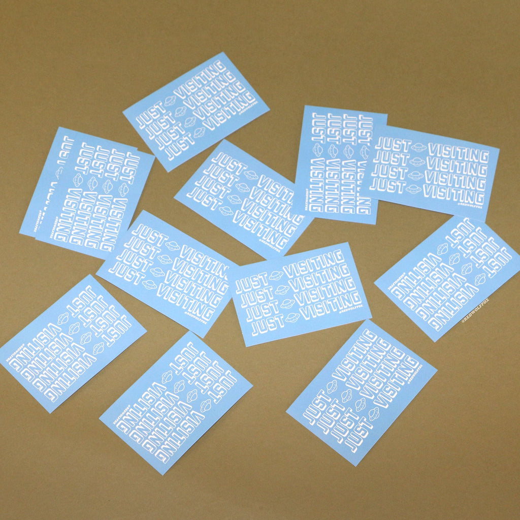 baby blue rectangle stickers with text 'JUST VISITING' printed across in white ink scattered across a vintage green backdrop