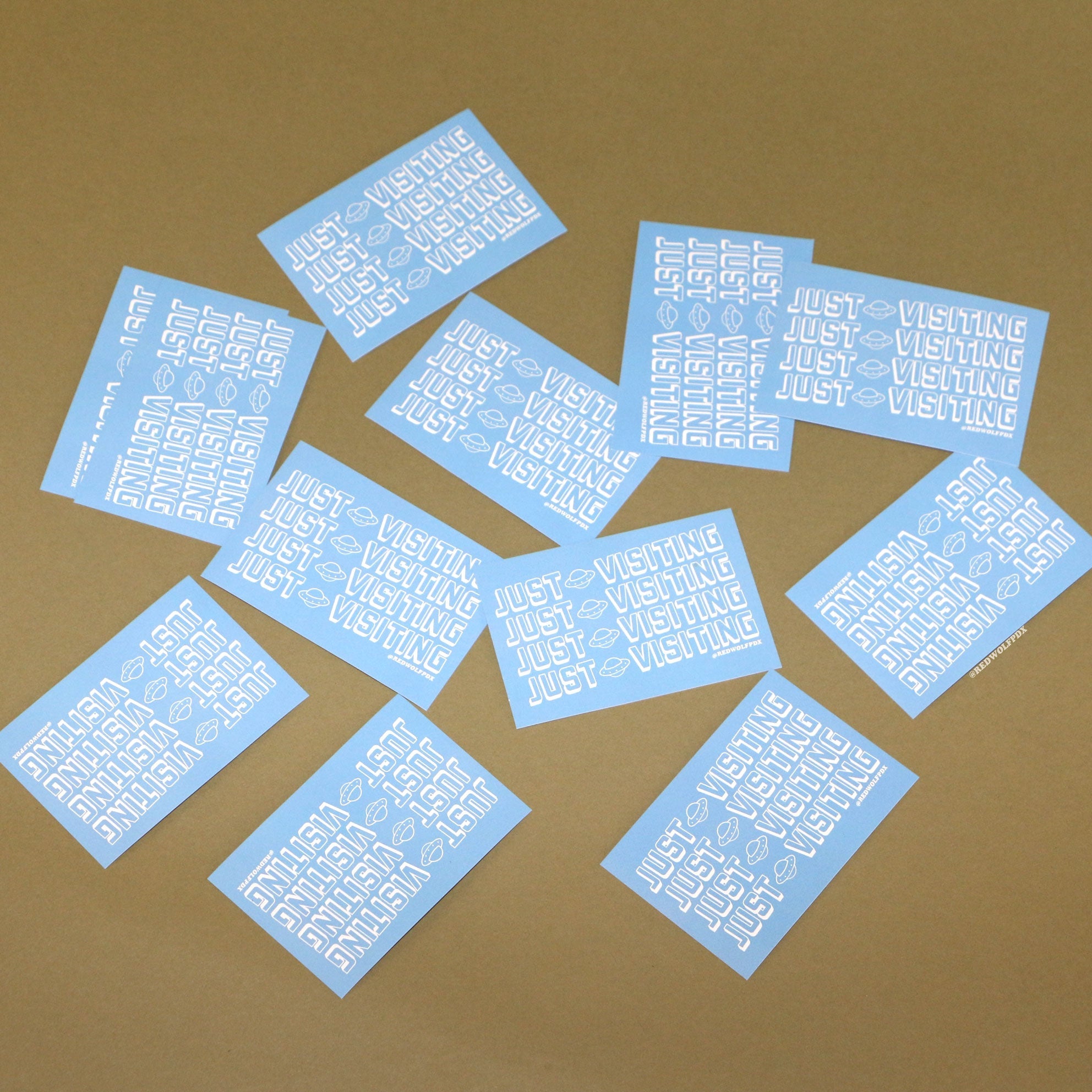 baby blue rectangle stickers with text 'JUST VISITING' printed across in white ink scattered across a vintage green backdrop
