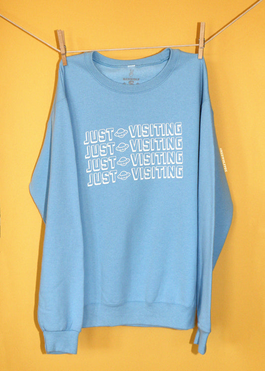 A light blue crewneck sweatshirt with 'JUST VISITING' printed in white across the chest, hanging from a clothesline with wooden clips against a solid yellow background.