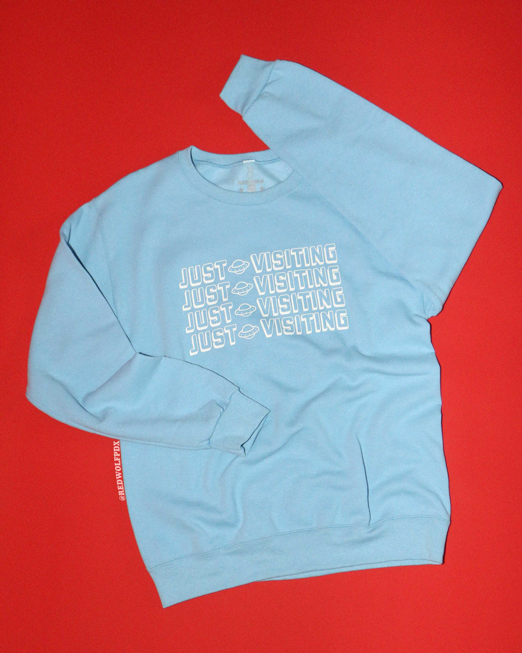 Baby blue crewneck sweatshirt with the text 'JUST VISITING' against red backdrop