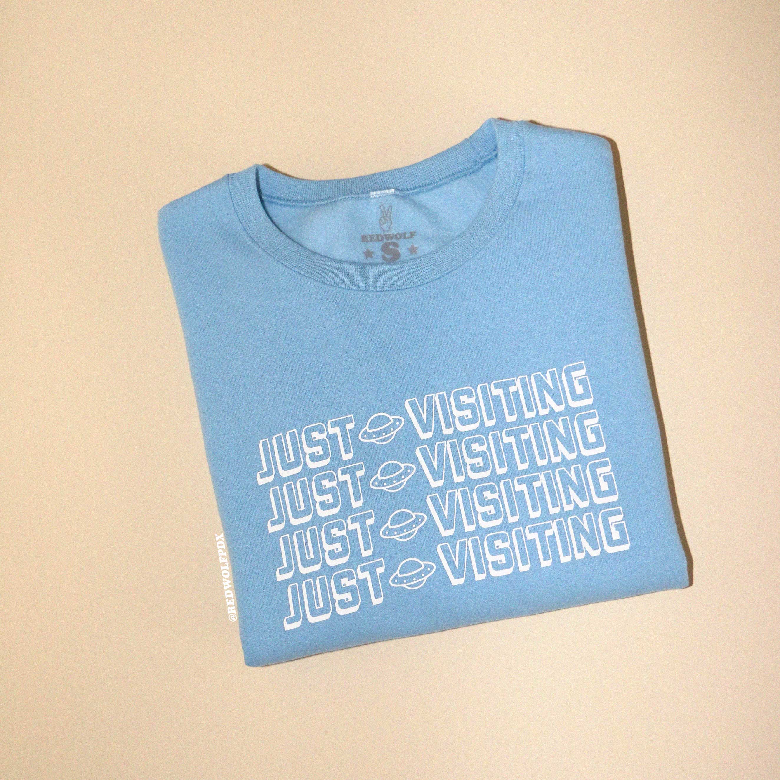 Folded baby blue crewneck sweatshirt with text 'JUST VISITING' on front against beige backdrop