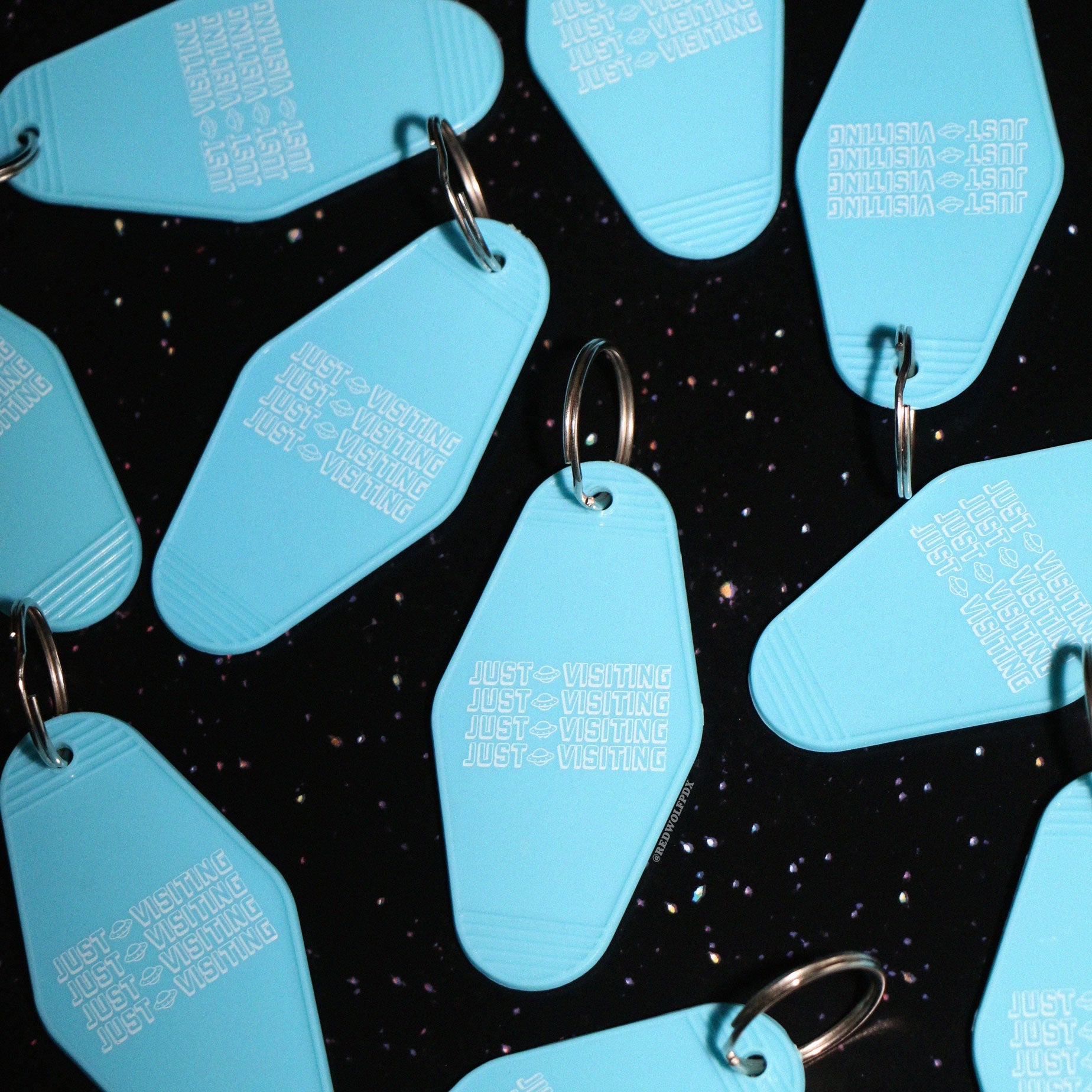 Baby blue retro motel style keychains with text 'JUST VISITING' printed across with white ink scattered on a space background