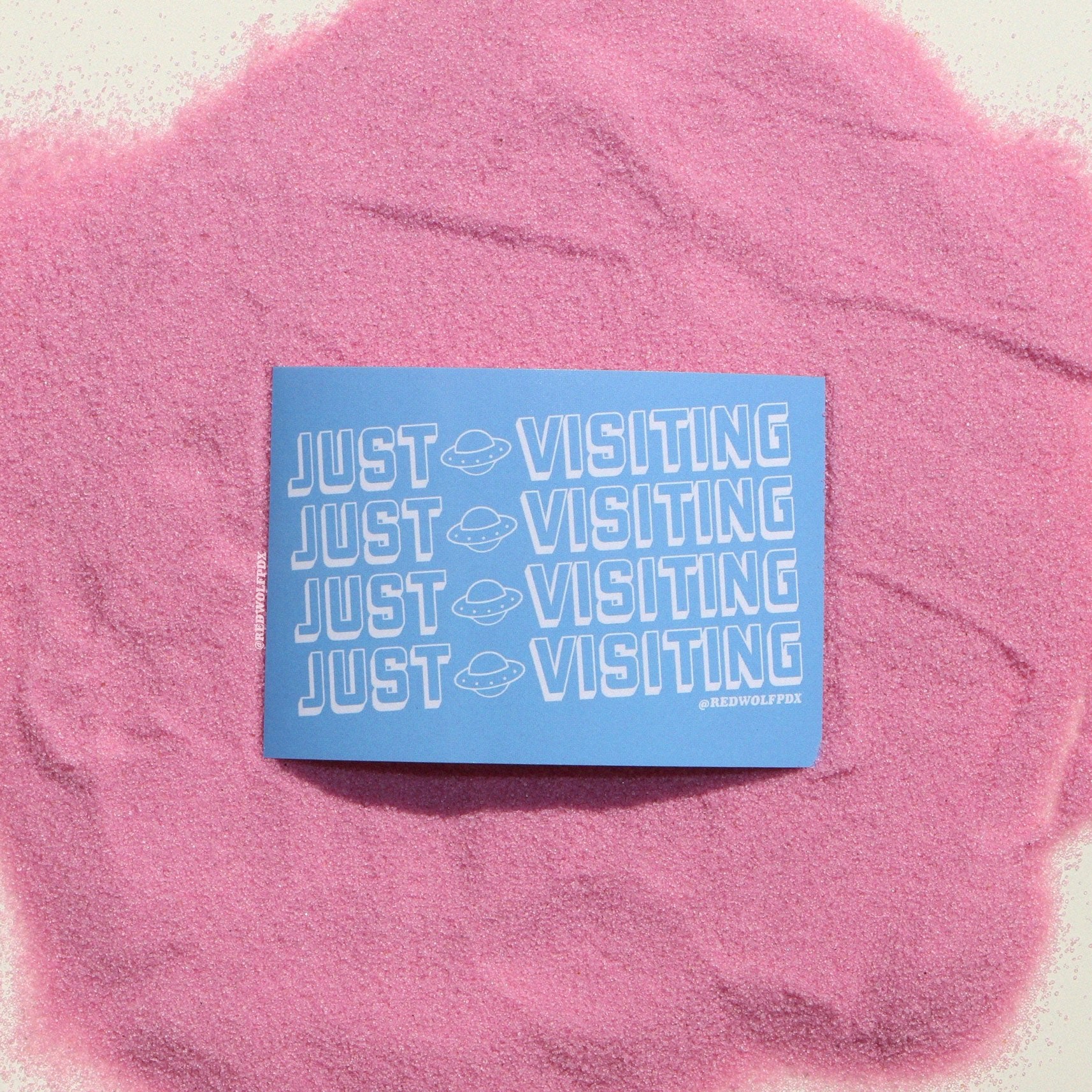baby blue rectangle sticker with text 'JUST VISITING' printed across in white rested on pink sand background