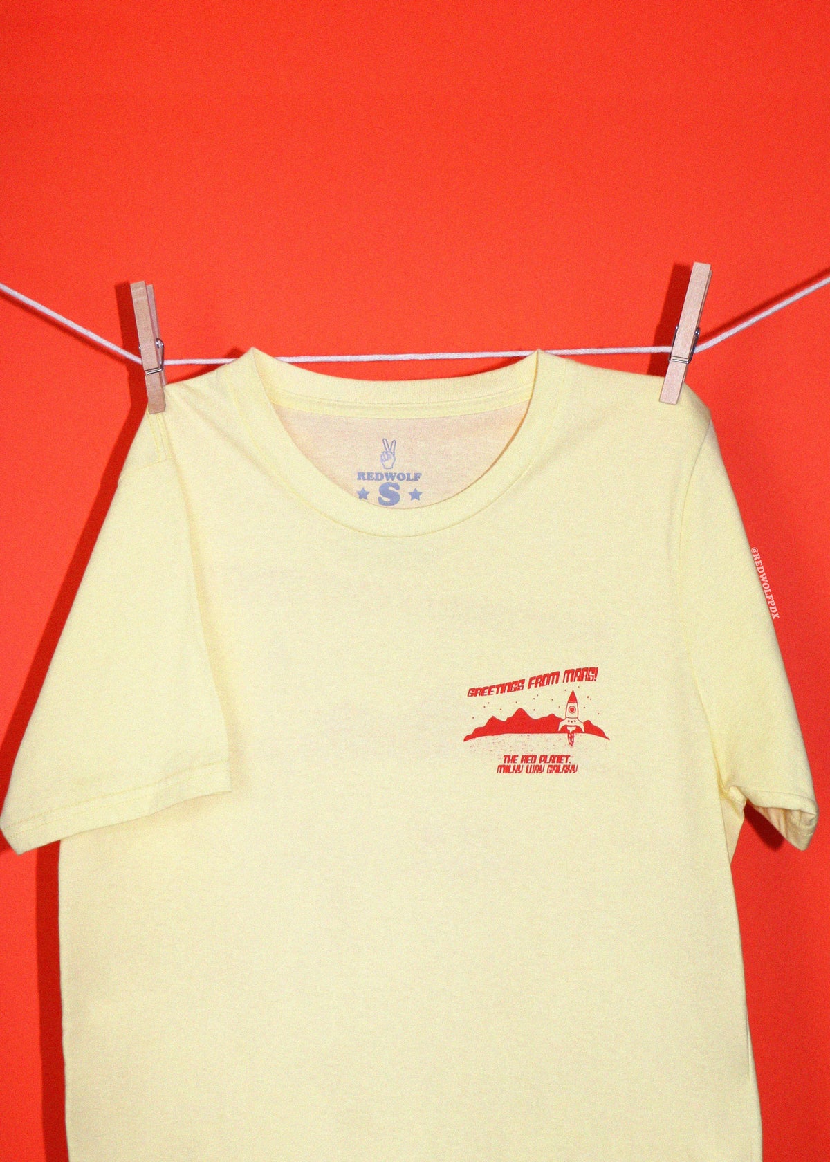A pale yellow short-sleeve t-shirt with a red graphic 'Greetings from Mars!' on the left chest, hanging from a clothesline with wooden clothespins against a solid red background.