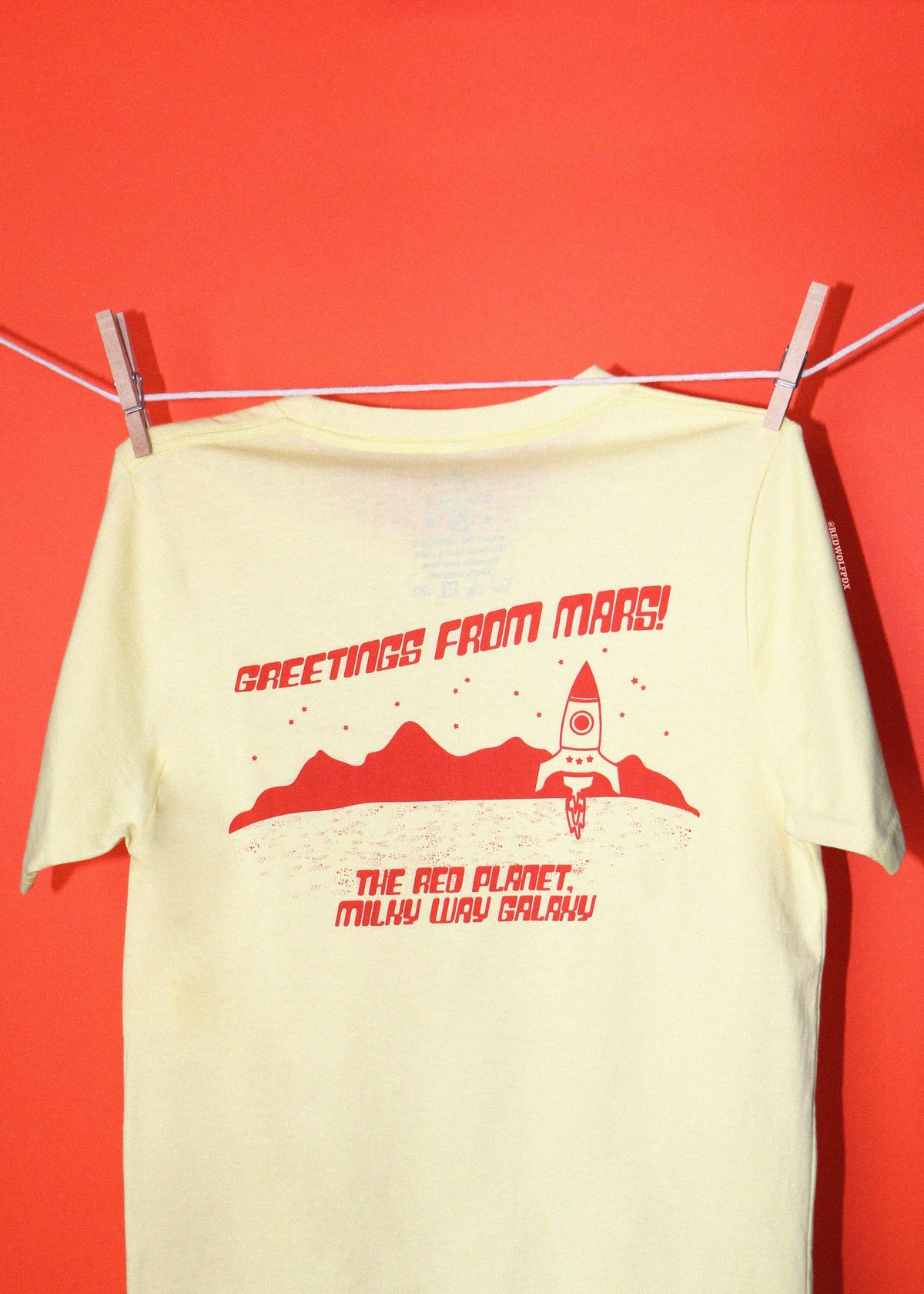 A pale yellow t-shirt hanging from a clothesline with wooden clothespins against a solid red-orange background. The shirt features a red graphic print of a rocket ship, mountains, and the text 'GREETINGS FROM MARS! THE RED PLANET.