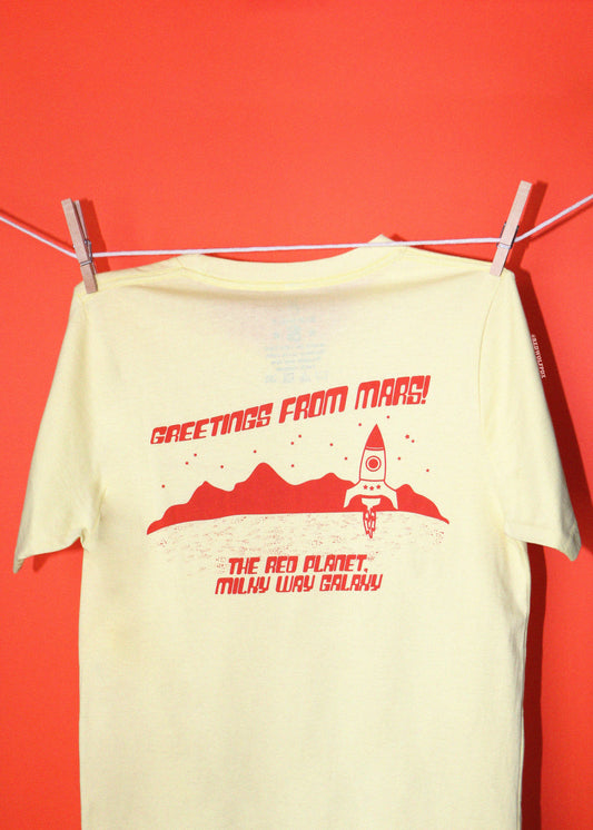 A pale yellow t-shirt hanging from a clothesline with wooden clothespins against a solid red-orange background. The shirt features a red graphic print of a rocket ship, mountains, and the text 'GREETINGS FROM MARS! THE RED PLANET.