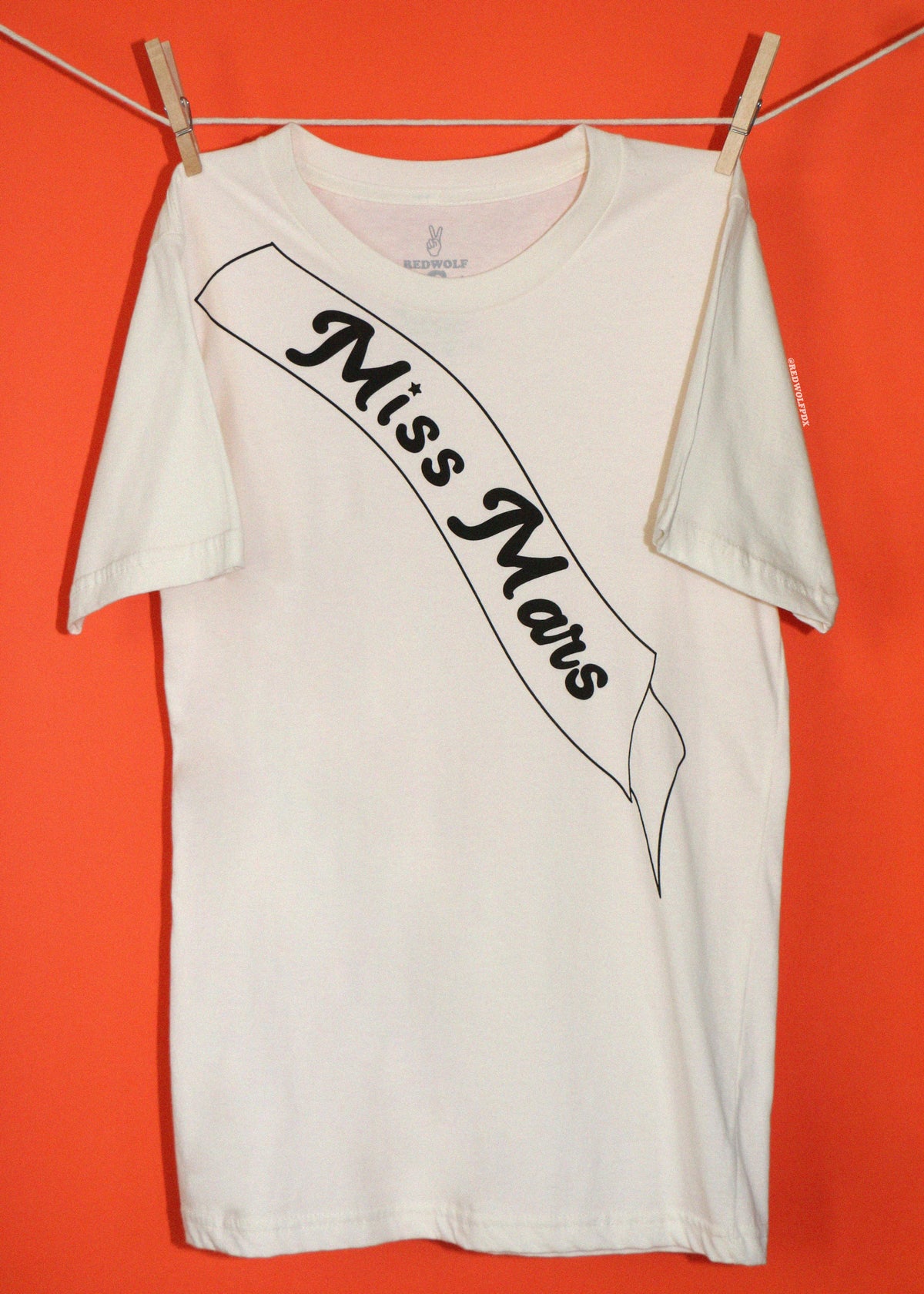 A white short-sleeve t-shirt hanging from a clothesline with wooden clothespins against a solid orange background. The shirt features a black graphic print of a banner reading 'Miss Mars' in a stylized font.