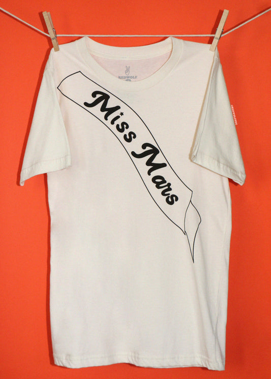 A white short-sleeve t-shirt hanging from a clothesline with wooden clothespins against a solid orange background. The shirt features a black graphic print of a banner reading 'Miss Mars' in a stylized font.