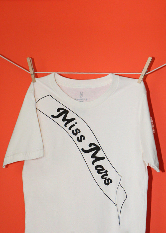 A white short-sleeve t-shirt with a black graphic print reading 'Miss Mars' on a sash design, hanging from wooden clothespins on a string against a solid orange background.