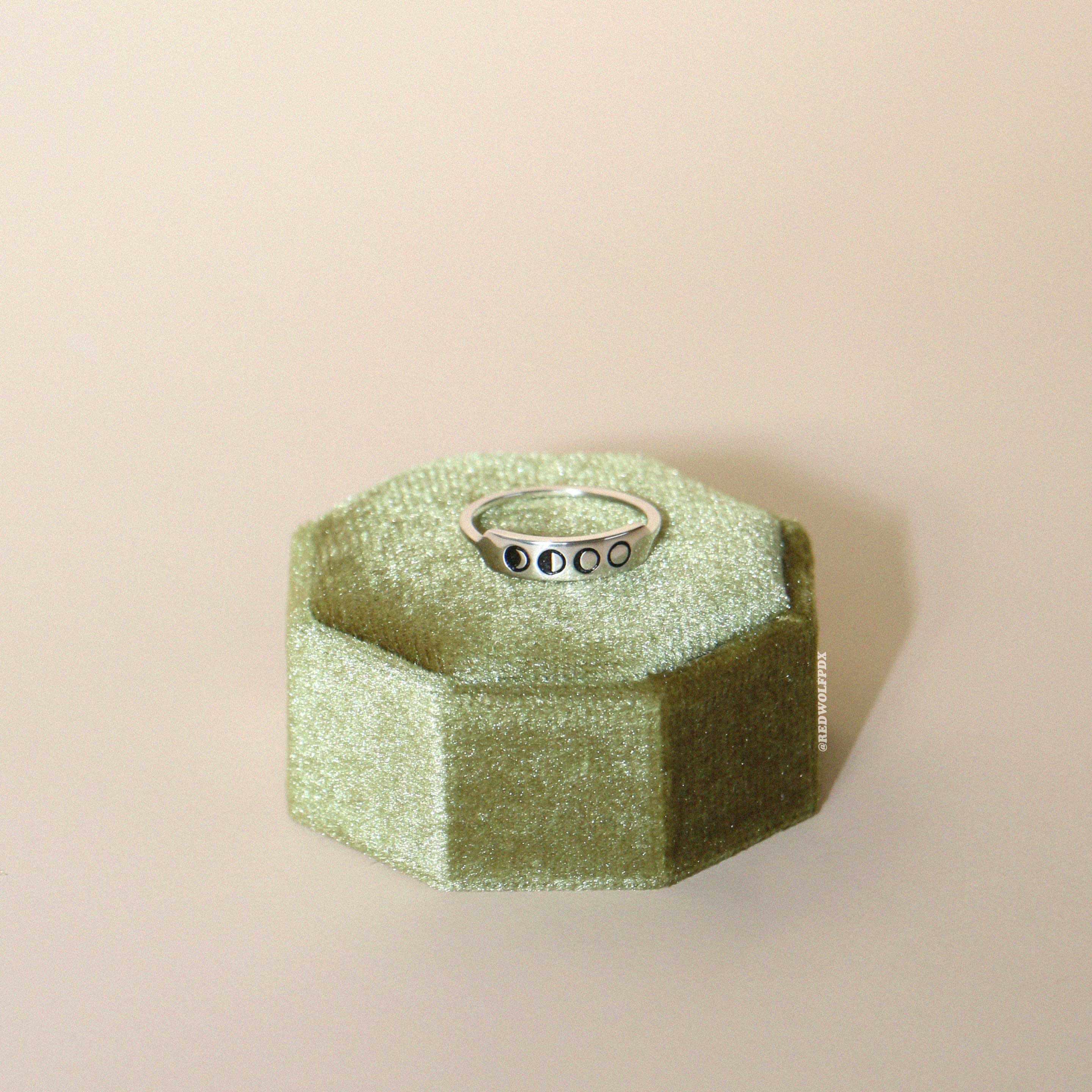 A sterling silver ring with four small moon phase engravings on it, displayed on a green velvet display on a beige background