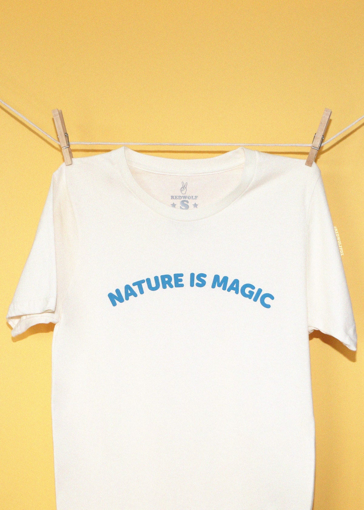 cream tee with text 'Nature is magic' screen printed across the chest in a bright blue hanging on a clothing line against a yellow backdrop.