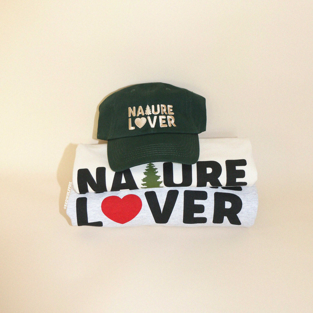 Green baseball cap with text 'NATURE LOVER' embroidered with beige thread across the front rested on stack of folded tee and sweatshirt both with text 'NATURE LOVER' screen printed across against beige backdrop