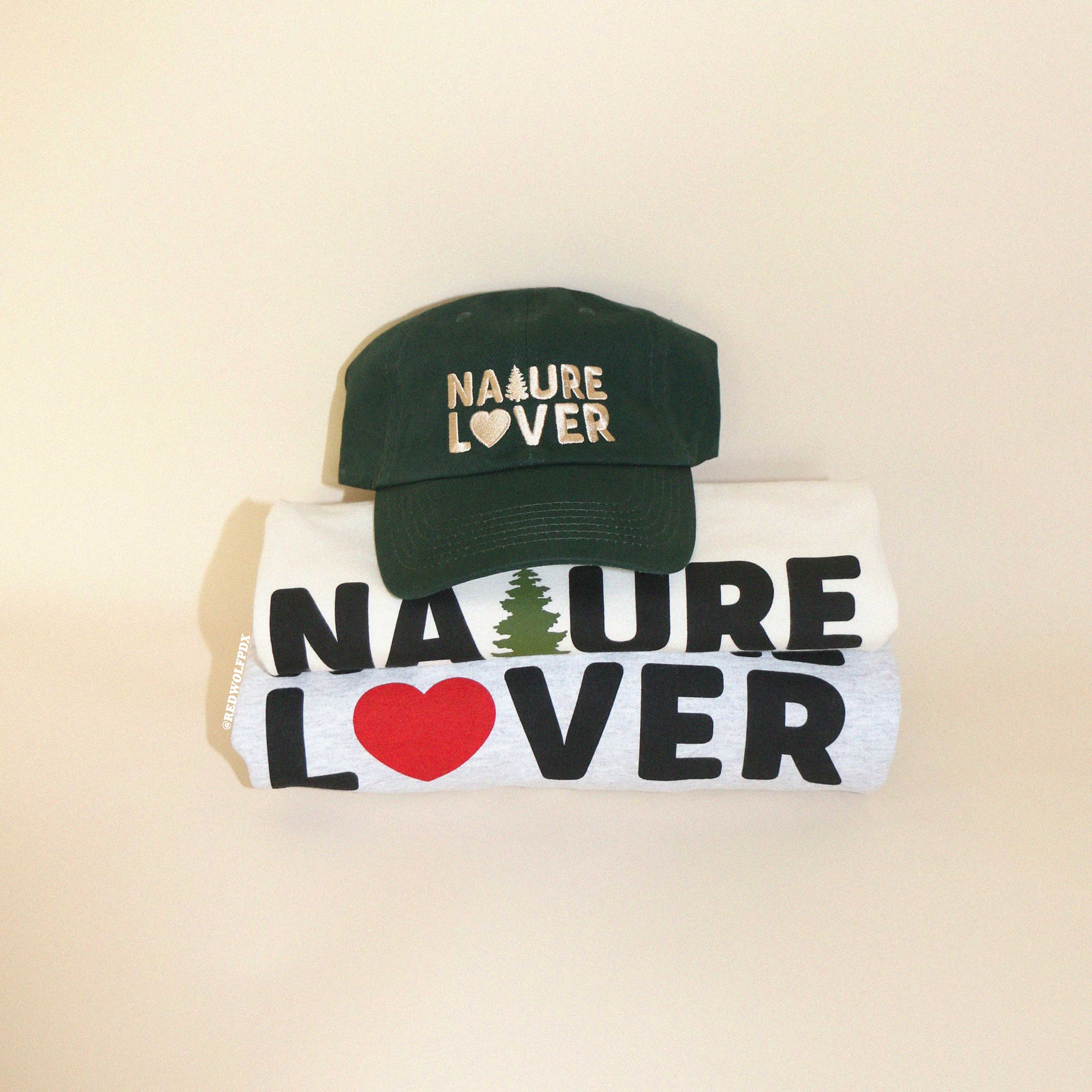 Green baseball cap with text 'NATURE LOVER' embroidered with beige thread across the front rested on stack of folded tee and sweatshirt both with text 'NATURE LOVER' screen printed across against beige backdrop