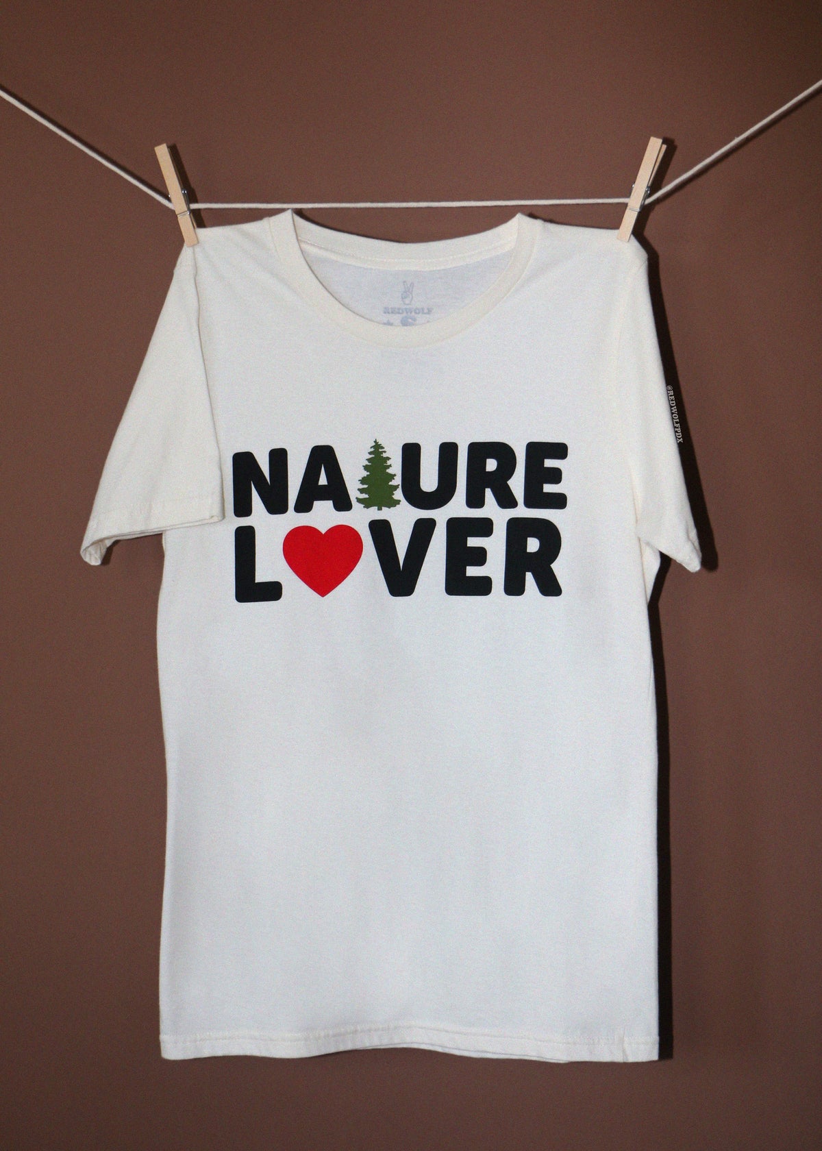 A white short-sleeve t-shirt hanging from a clothesline with wooden clothespins against a solid brown background. The shirt features a graphic print that reads 'NATURE LOVER' in black block letters, with a small green pine tree representing the letter 'A' and a red heart representing the letter 'O'.