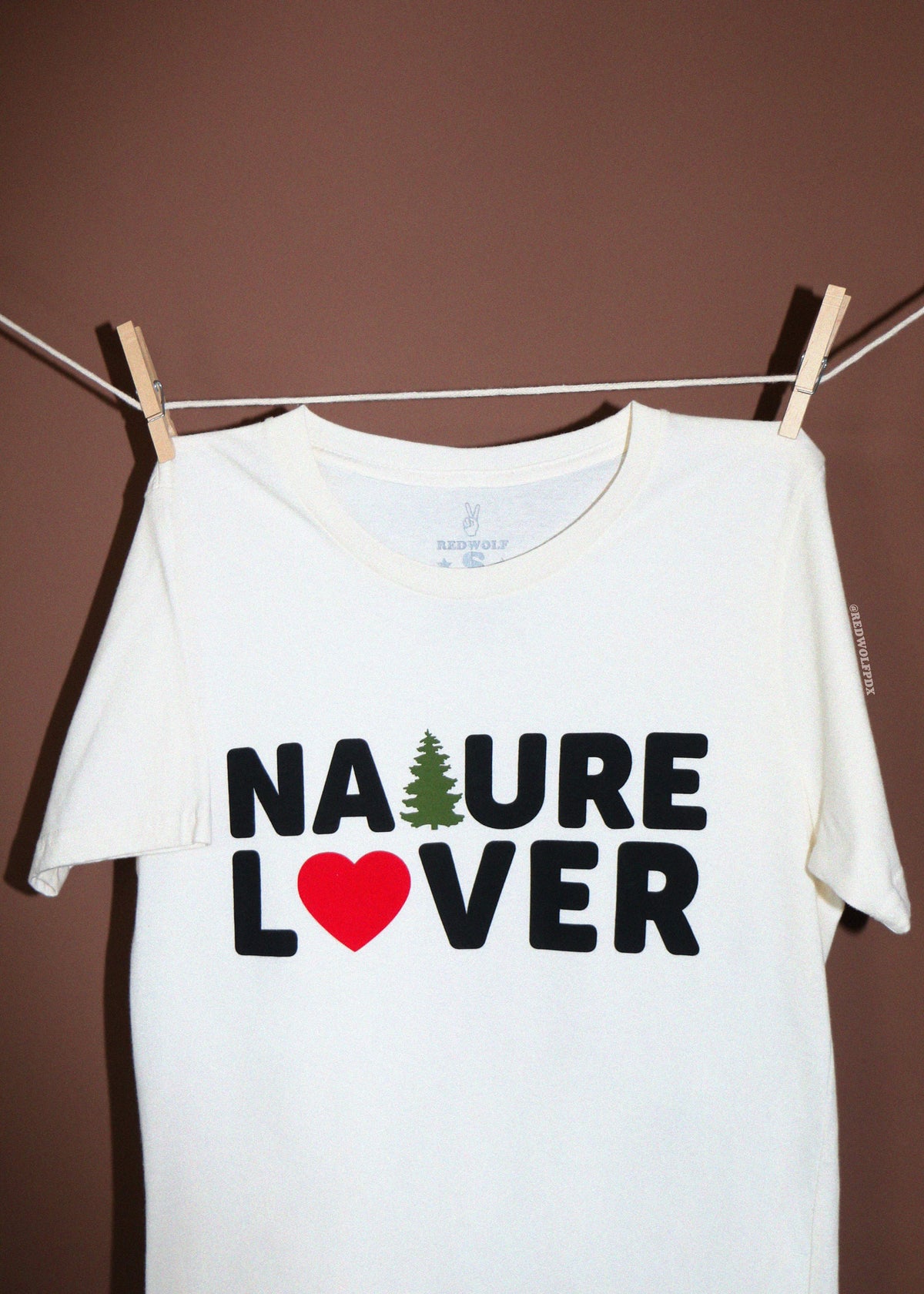 A white short-sleeve t-shirt hanging from a clothesline with wooden clothespins against a solid brown wall. The shirt features a graphic print that reads 'NATURE LOVER' in black letters, with a green pine tree representing the letter 'A' and a red heart representing the letter 'O'.