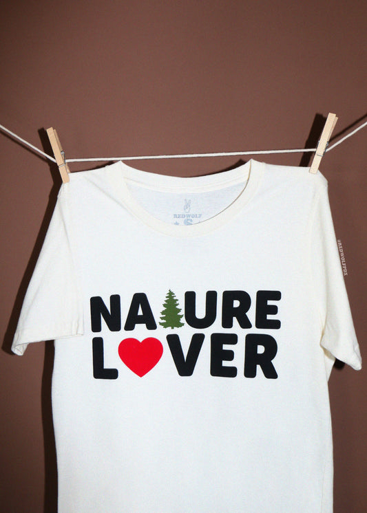 A white short-sleeve t-shirt hanging from a clothesline with wooden clothespins against a solid brown wall. The shirt features a graphic print that reads 'NATURE LOVER' in black letters, with a green pine tree representing the letter 'A' and a red heart representing the letter 'O'.