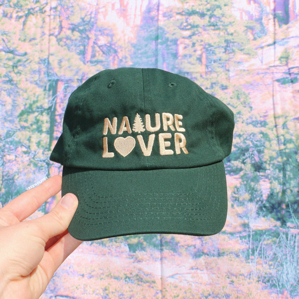 forest green baseball cap with text 'NATURE LOVER' embroidered in beige thread across front being held against a nature backdrop