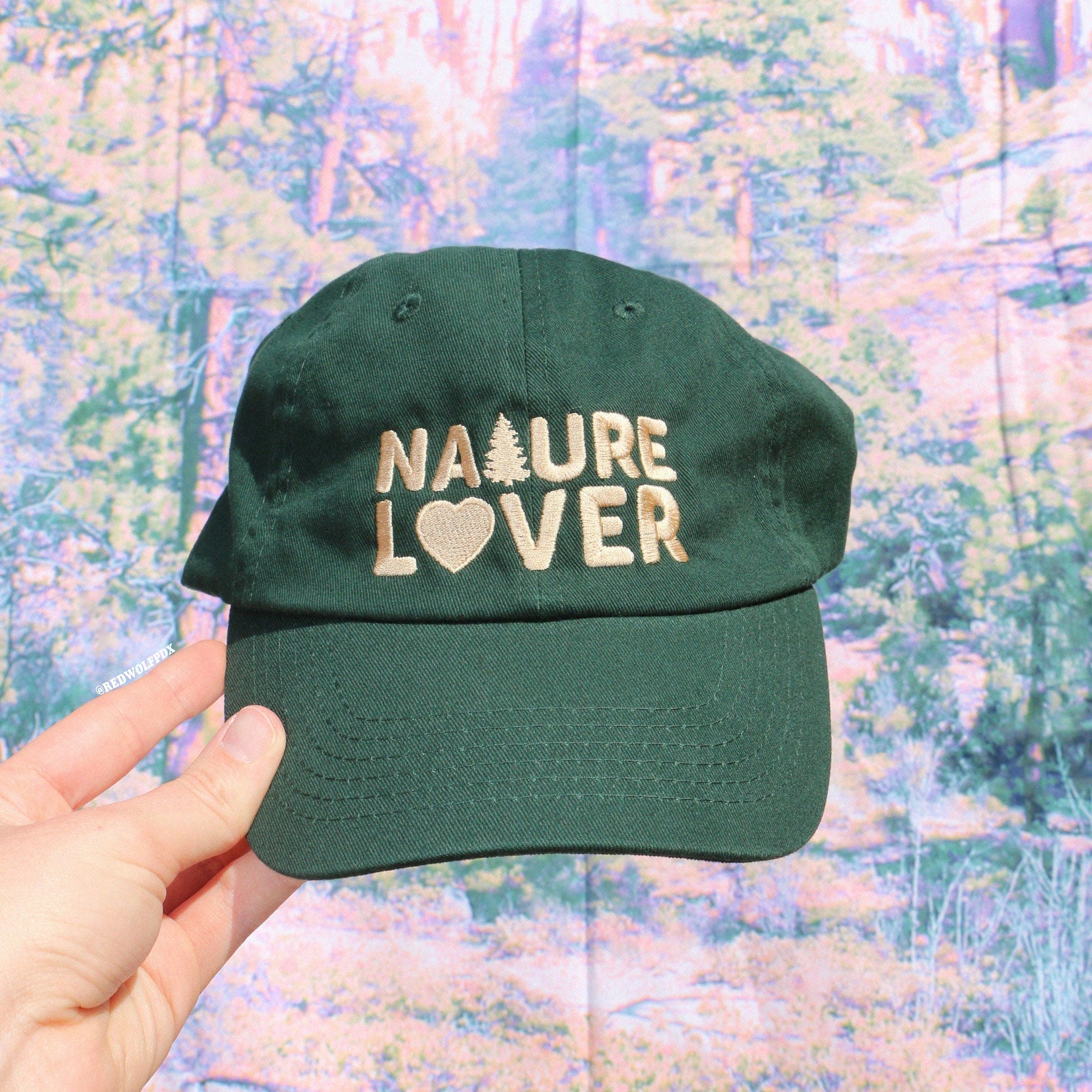 forest green baseball cap with text 'NATURE LOVER' embroidered in beige thread across front being held against a nature backdrop