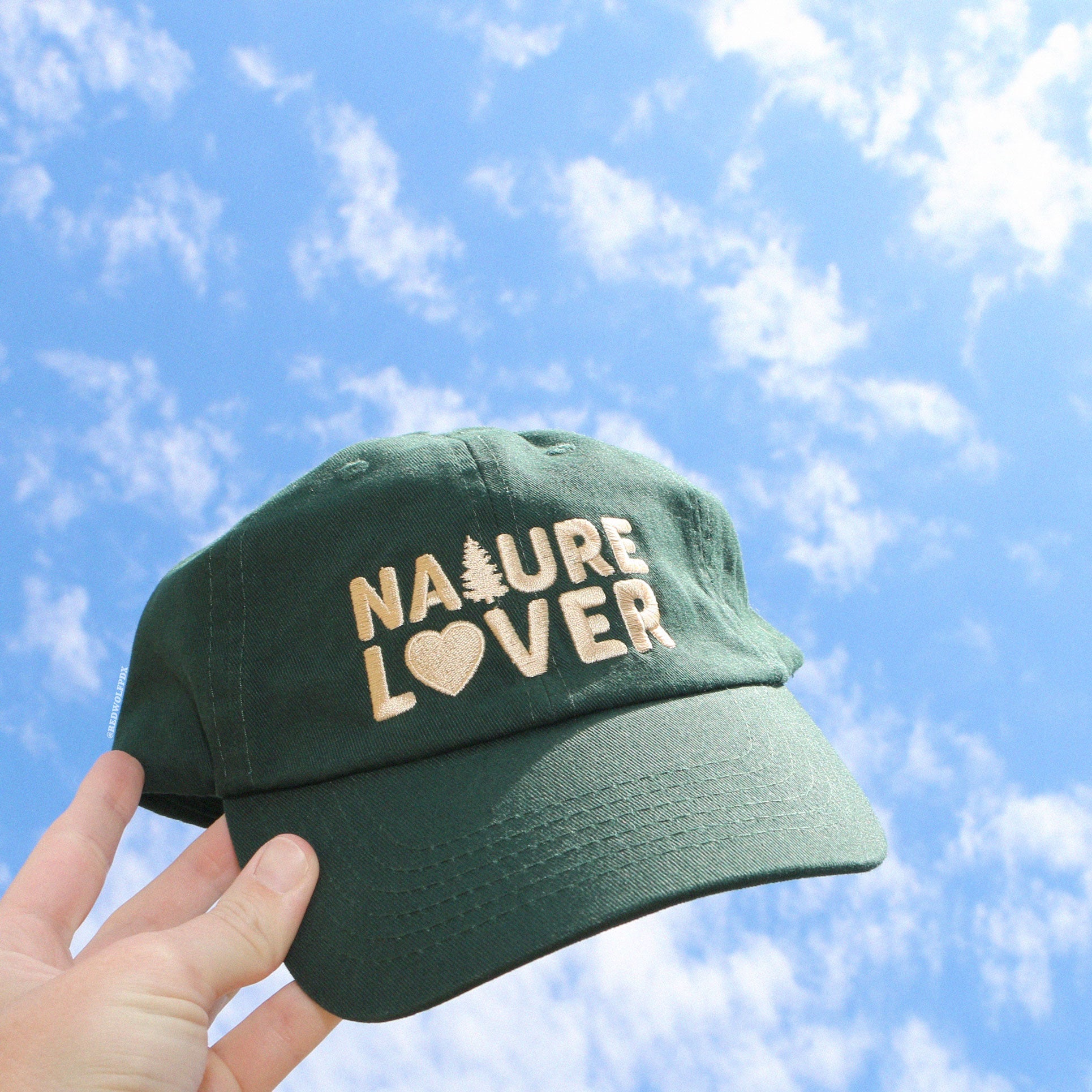 a forest green baseball cap with text 'NATURE LOVER' embroidered with beige thread across the front being held up by a hand to blue sky with fluffy white clouds
