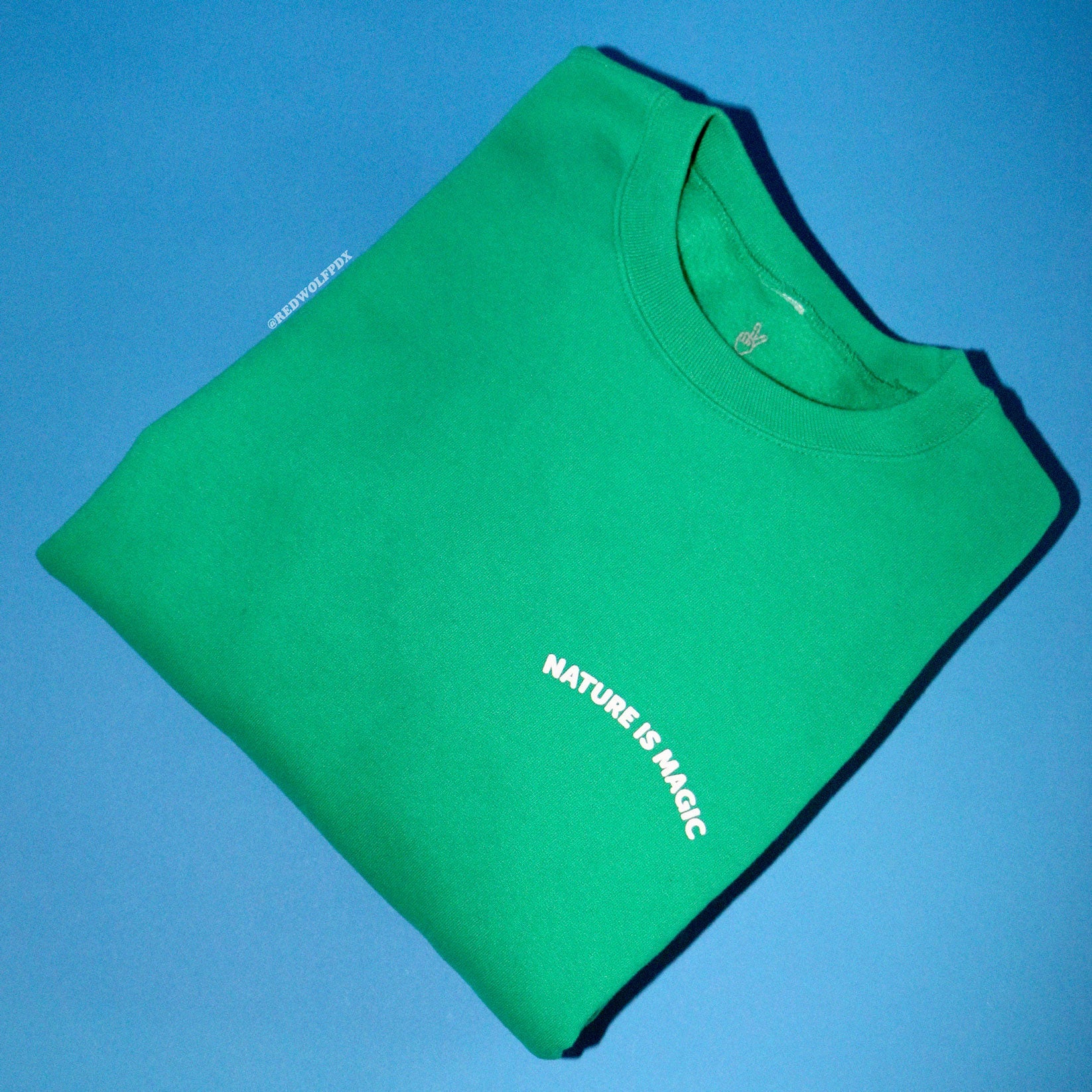 A folded green crewneck sweatshirt with the text 'NATURE IS MAGIC' screen printed on the front.