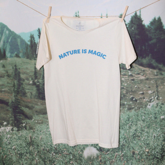 beige graphic tee with text 'NATURE IS MAGIC' in blue ink screen printed across chest hanging from clothing line against a nature backdrop