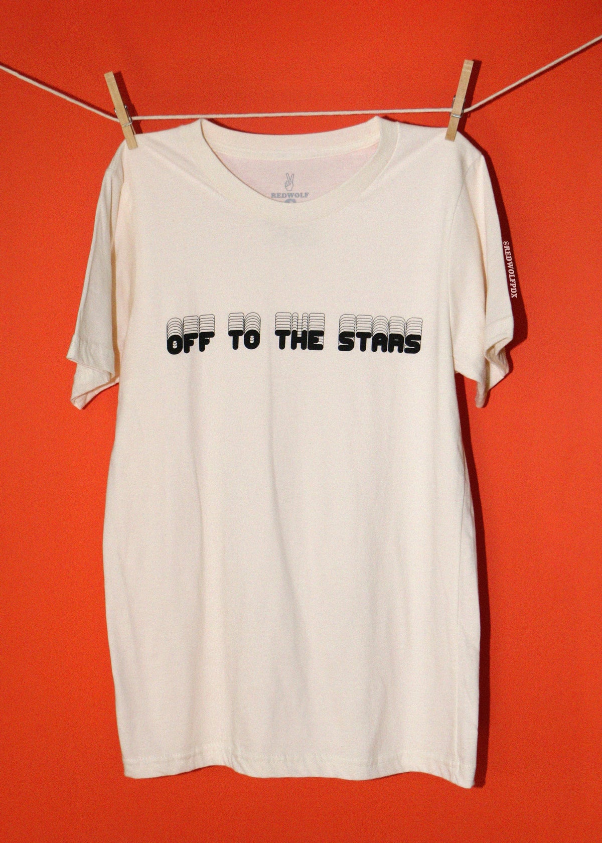 An off-white short-sleeve t-shirt hanging from a clothesline with wooden clips against a solid orange-red background. The shirt features the text 'OFF TO THE STARS' printed across the chest in black, with small gray rectangular graphics preceding each word.