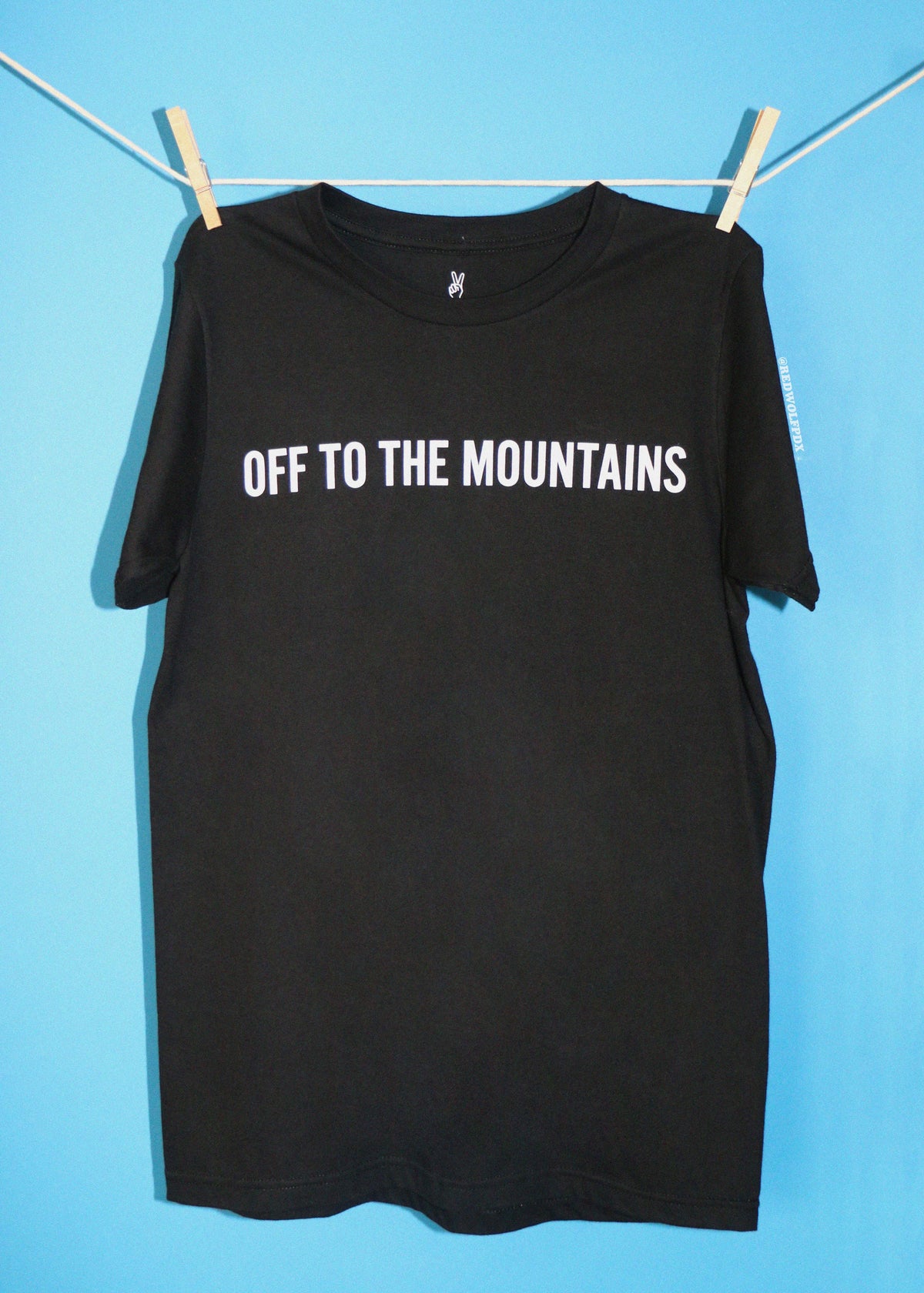 A black short-sleeve t-shirt with 'OFF TO THE MOUNTAINS' printed in white across the chest, hanging from a clothesline with wooden clothespins against a bright blue background.