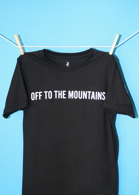 A black short-sleeve t-shirt with 'OFF TO THE MOUNTAINS' printed in white across the chest, hanging from a white clothesline with wooden clothespins against a solid light blue background.