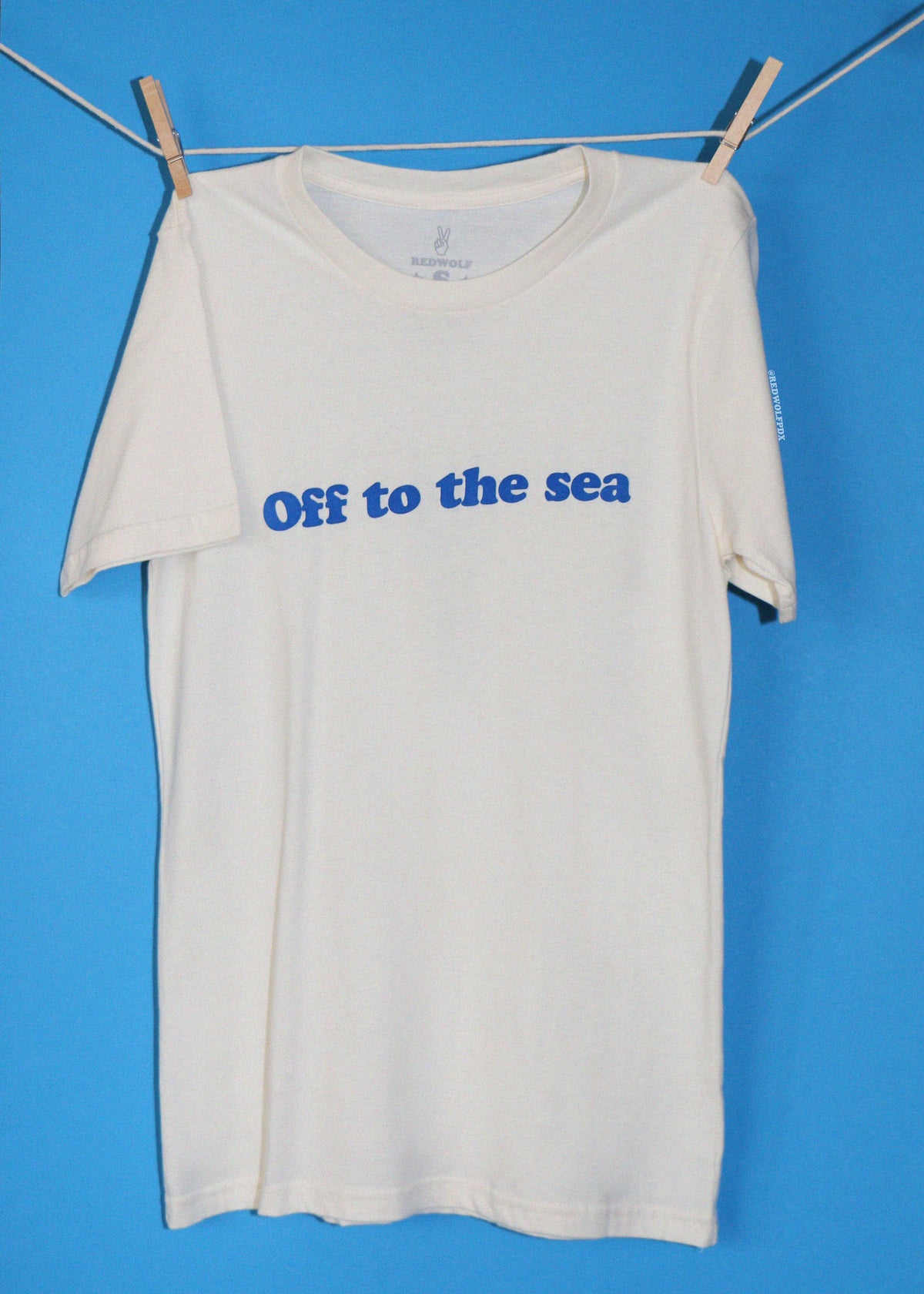 An off-white short-sleeve t-shirt with 'Off to the sea' printed in blue across the chest, hanging from a clothesline with wooden clothespins against a solid blue background.