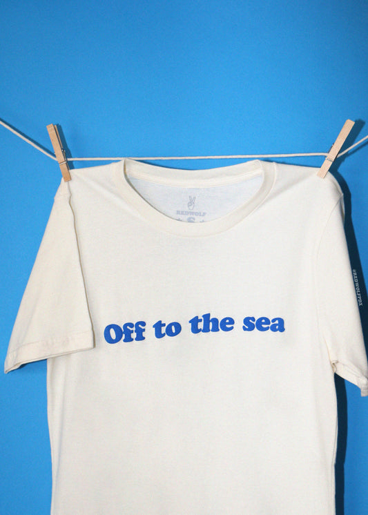 An off-white short-sleeve t-shirt with 'Off to the sea' printed in blue across the chest, hanging from a clothesline with wooden clothespins against a solid blue background.
