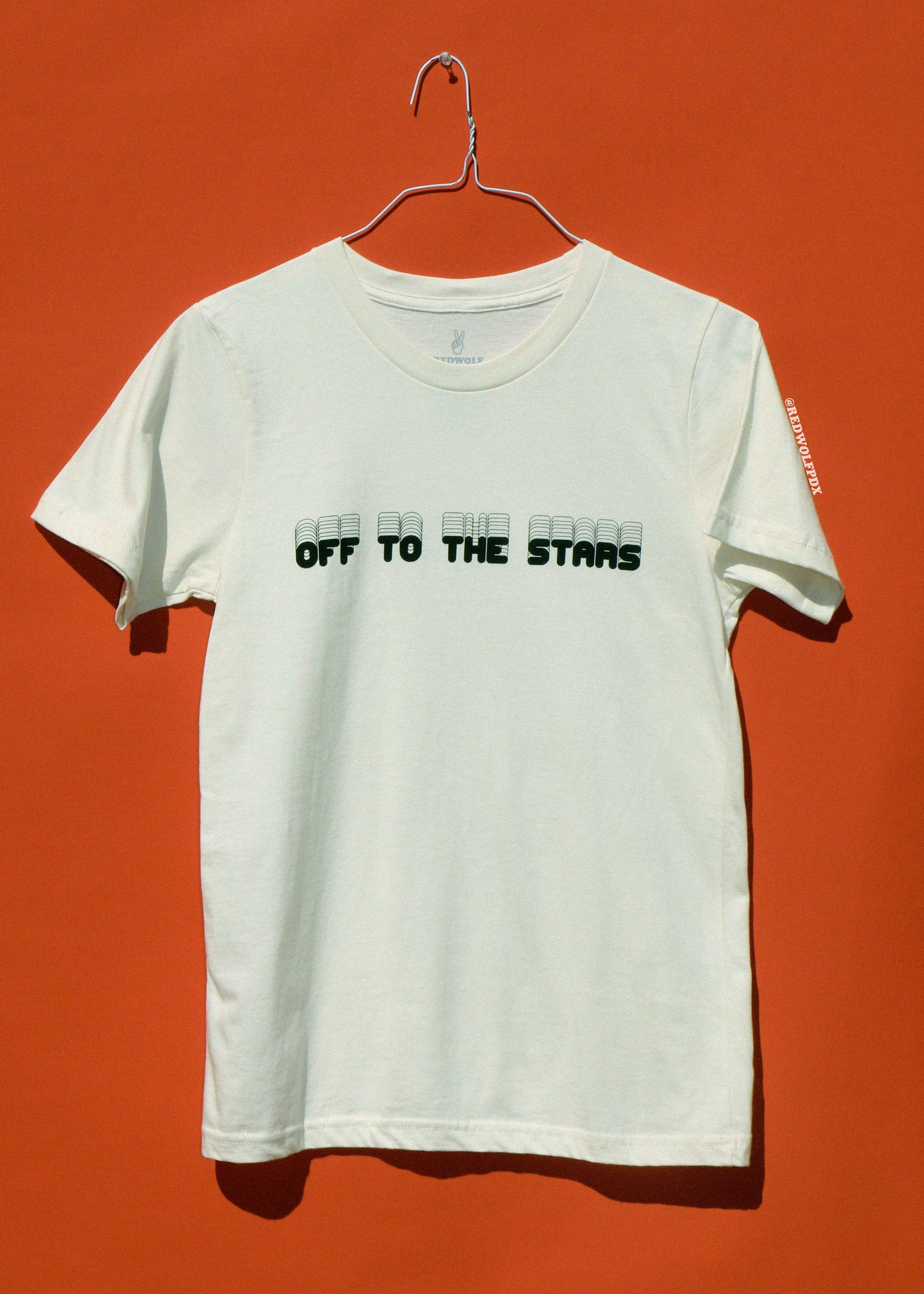 White t-shirt with text 'Off to the Stars' on a hanger against an orange background