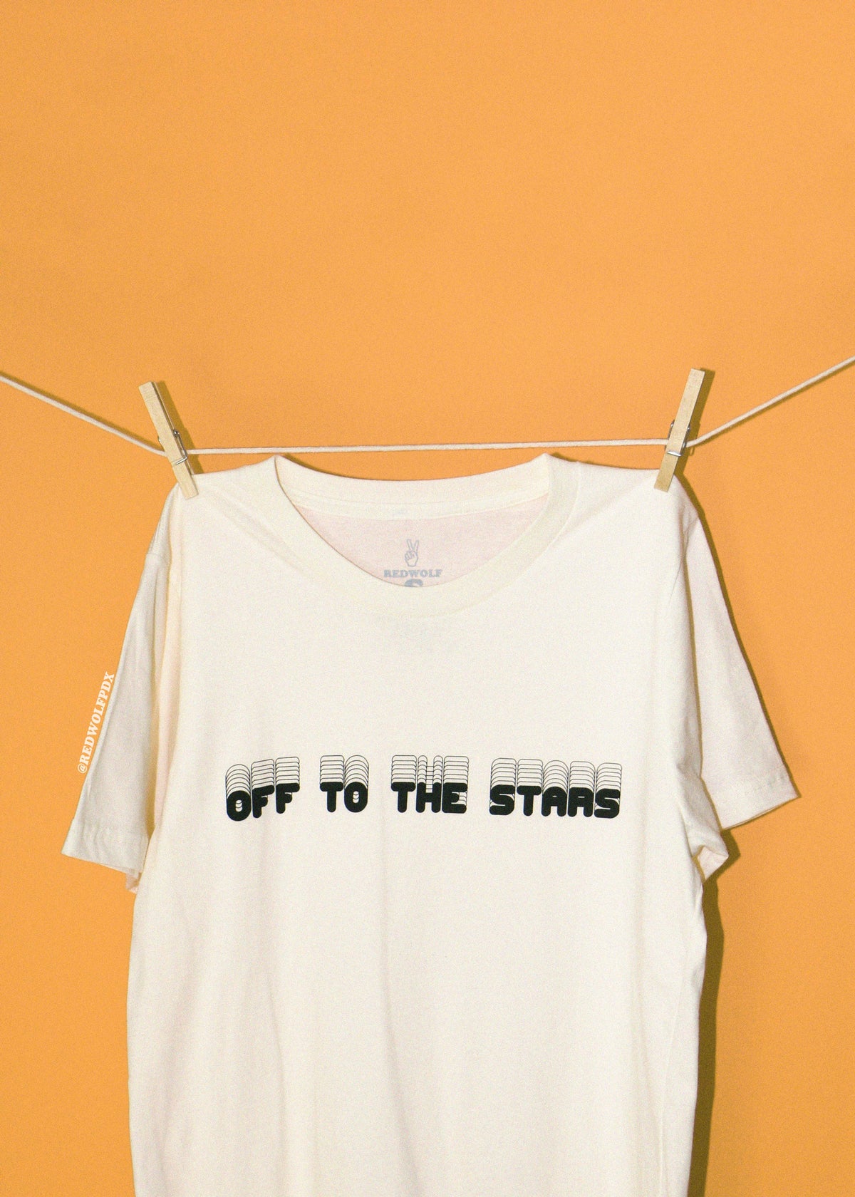 An off-white short-sleeve t-shirt with 'OFF TO THE STARS' printed in black across the chest, hanging from a clothesline with wooden clips against a solid golden yellow background.