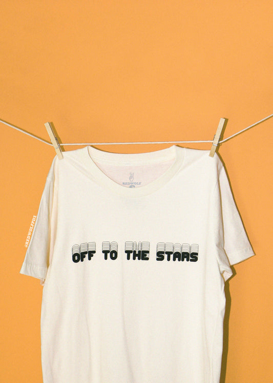 An off-white short-sleeve t-shirt with 'OFF TO THE STARS' printed in black across the chest, hanging from a clothesline with wooden clips against a solid golden yellow background.