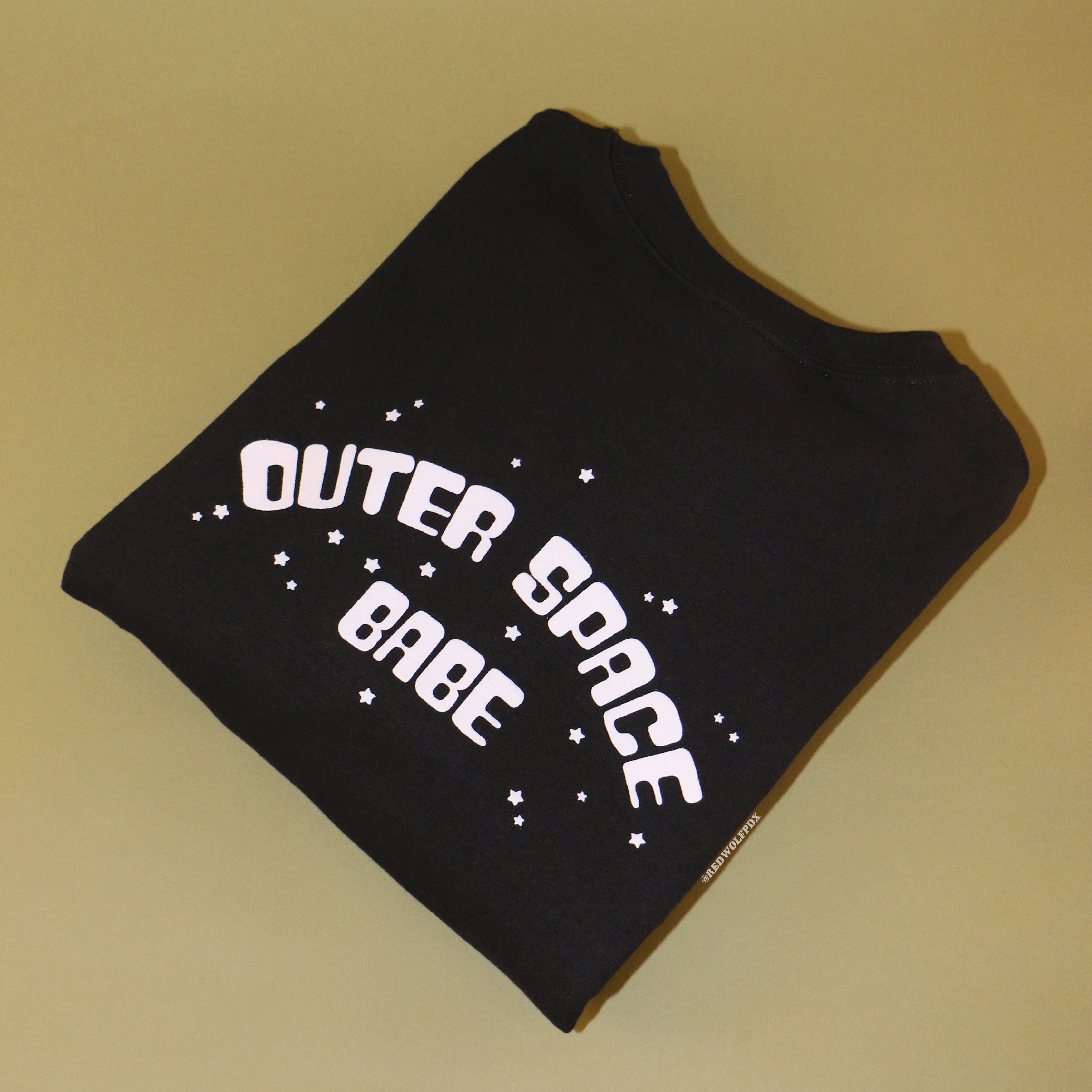 Folded black crewneck sweatshirt with text 'OUTER SPACE BABE' pink ink screen printed across the back on a vintage green background