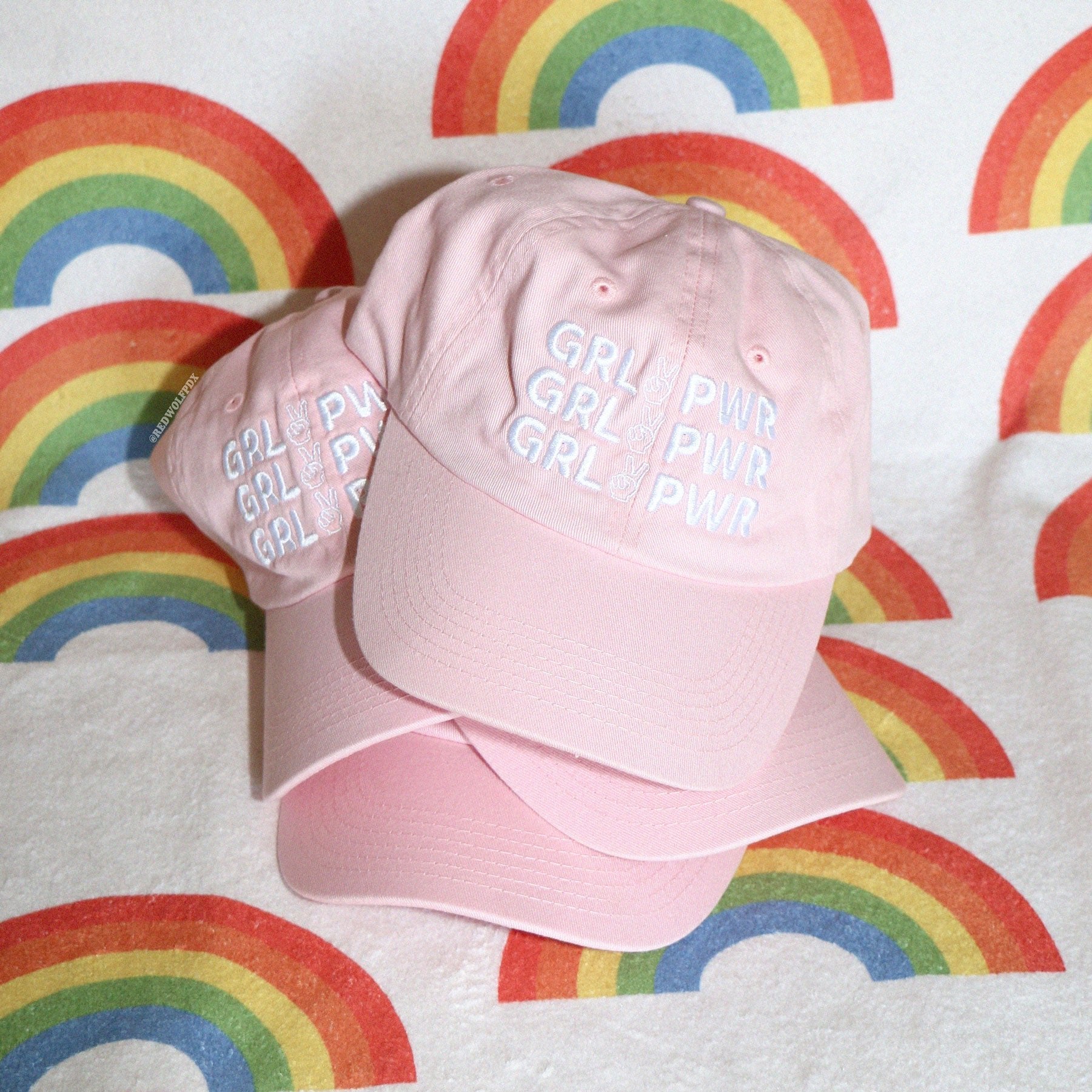 Stack of pink baseball caps with text 'GRL PWR' embroidered across with white thread on a retro rainbow towel backdrop