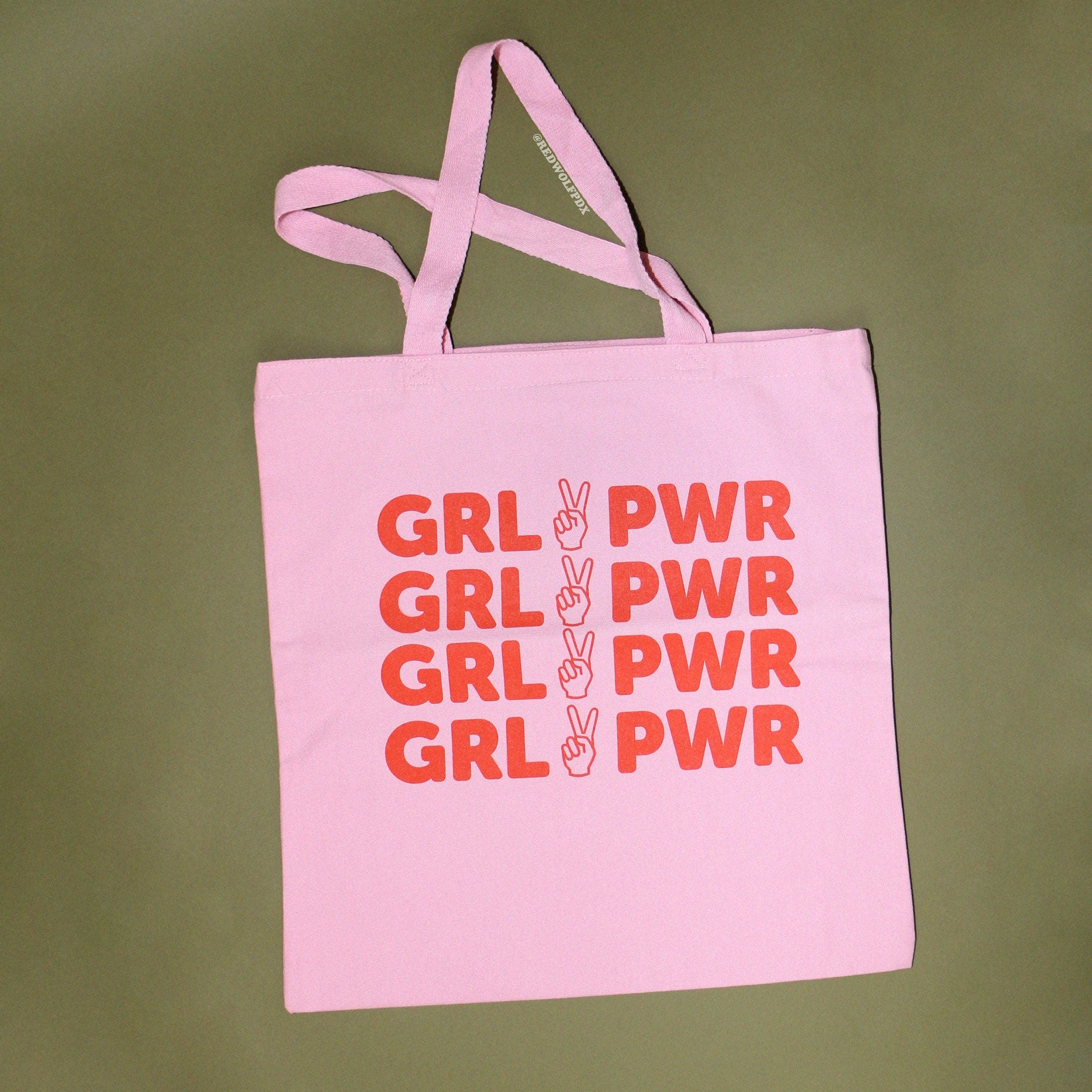 light pink canvas tote bag with text 'GRL PWR' in orange red ink screen printed down the front against a vintage green backdrop