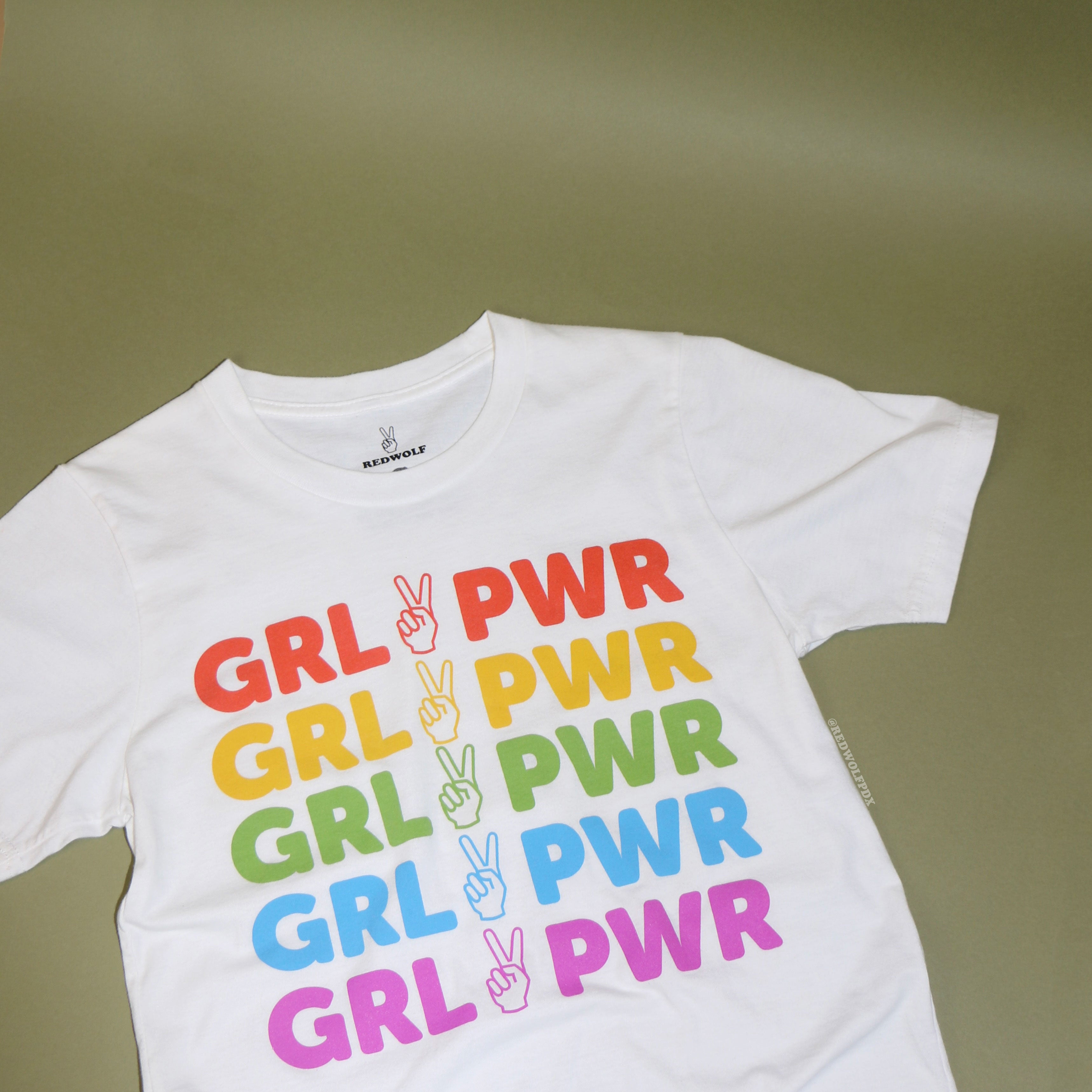 white tee with text 'GRL PWR' with peace sign graphic screen printed in rainbow colors down the front against a green background