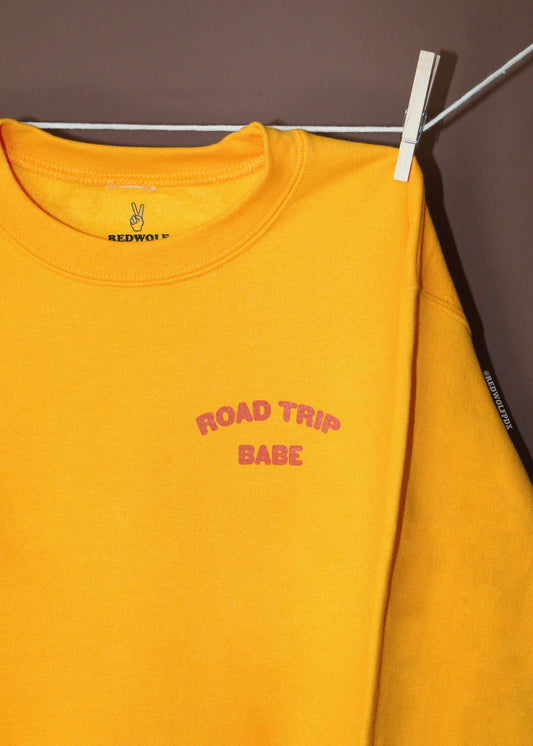 A bright yellow crewneck sweatshirt hanging from a white clothespin on a metal hanger against a dark brown wall. The sweatshirt features 'ROAD TRIP BABE' printed in pink arched text on the left chest, and the brand name 'REDWOLF' is visible on the inner neck label and sleeve.