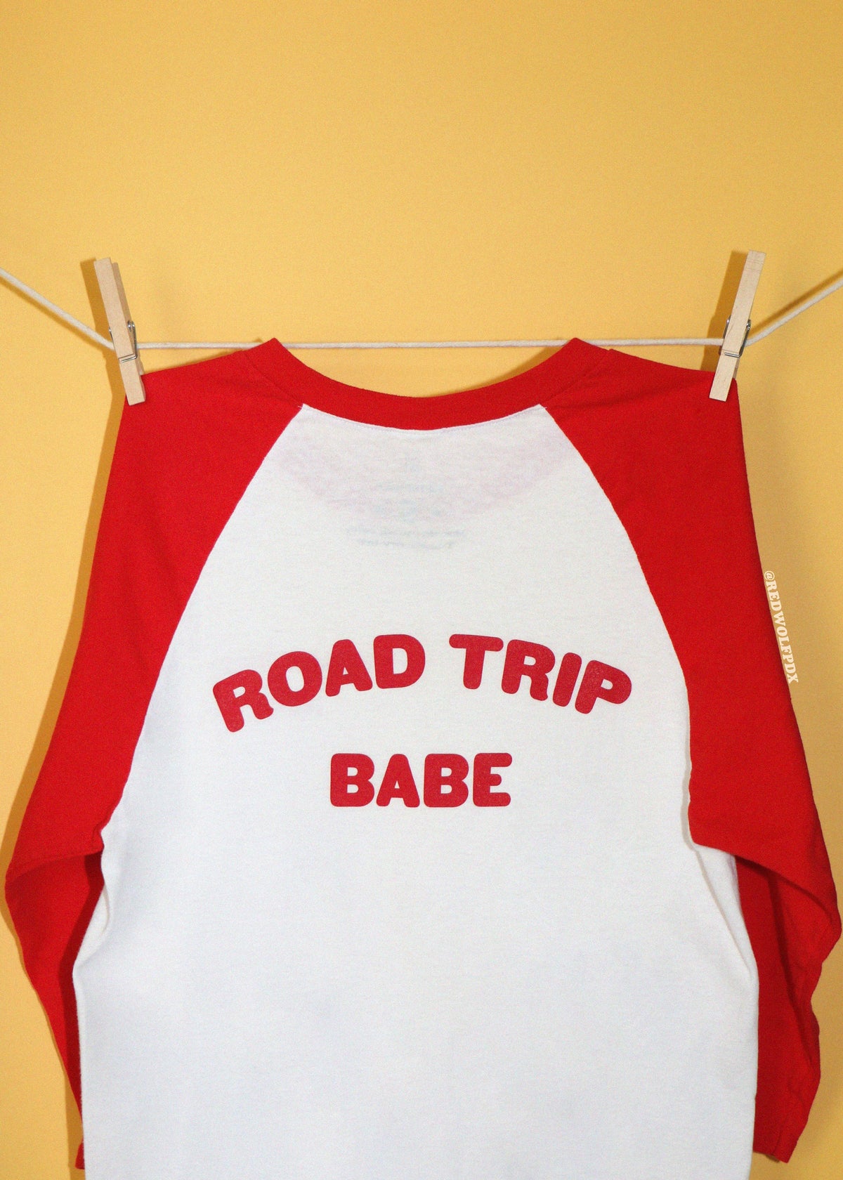 A red and white raglan baseball t-shirt hanging from a clothesline with wooden clothespins against a solid yellow background. The front of the shirt features the text 'ROAD TRIP BABE' printed in red arched letters.