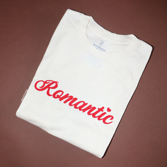 Folded beige graphic tee with text 'Romantic' in red ink screen printed across front on a brown backdrop