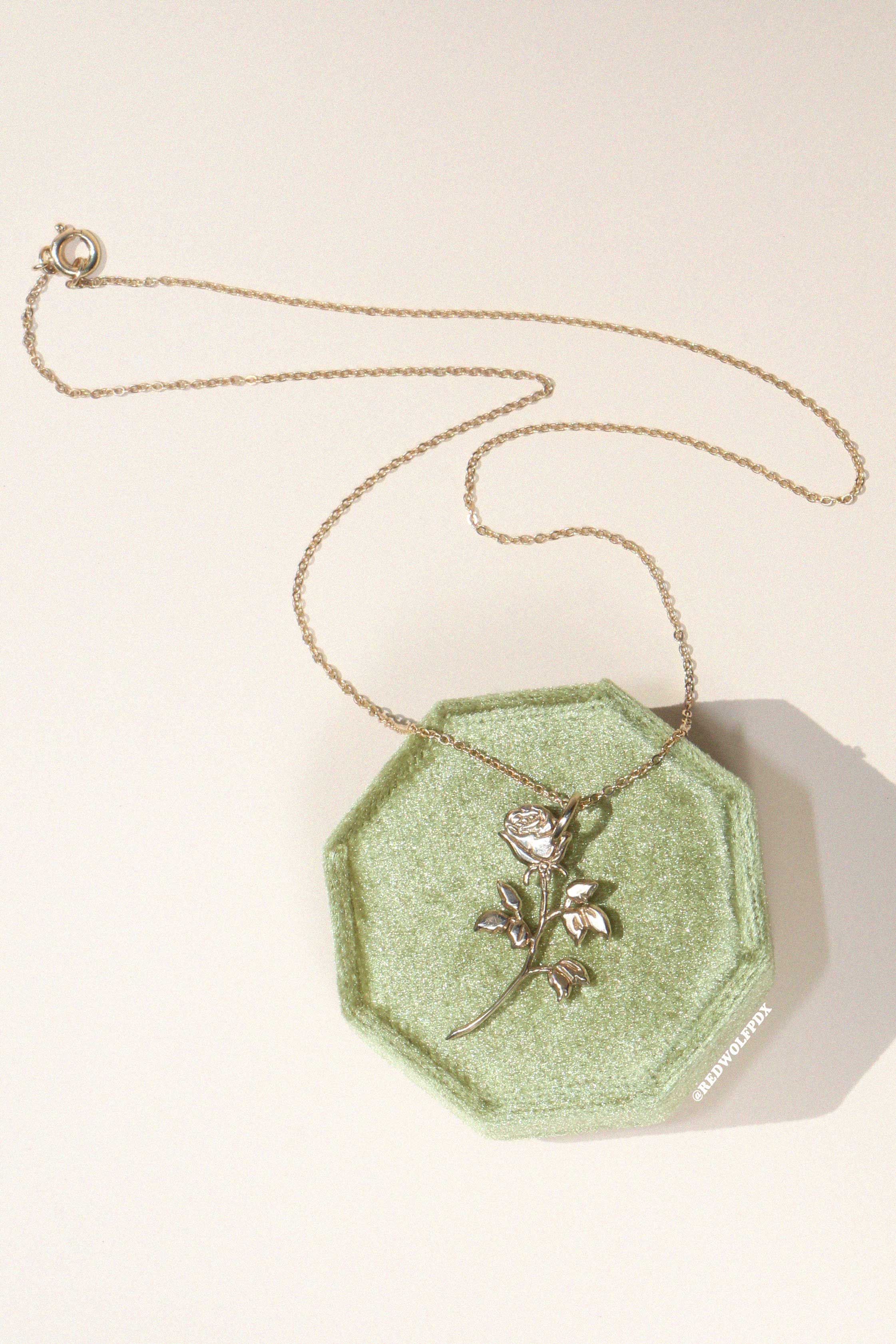 Rose charm in brass on a brass chain displayed on a vintage green jewelry box on beige backdrop