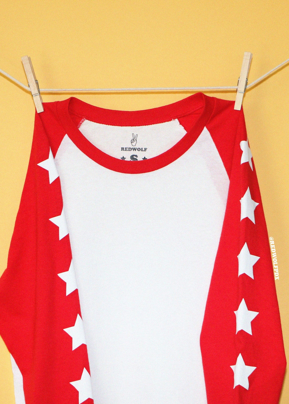 A red and white raglan long-sleeve shirt with white stars printed down the sleeves, hanging from a wooden clothesline with wooden clothespins against a solid yellow background. The brand name 'REDWOLF' is visible on the inner neck label.