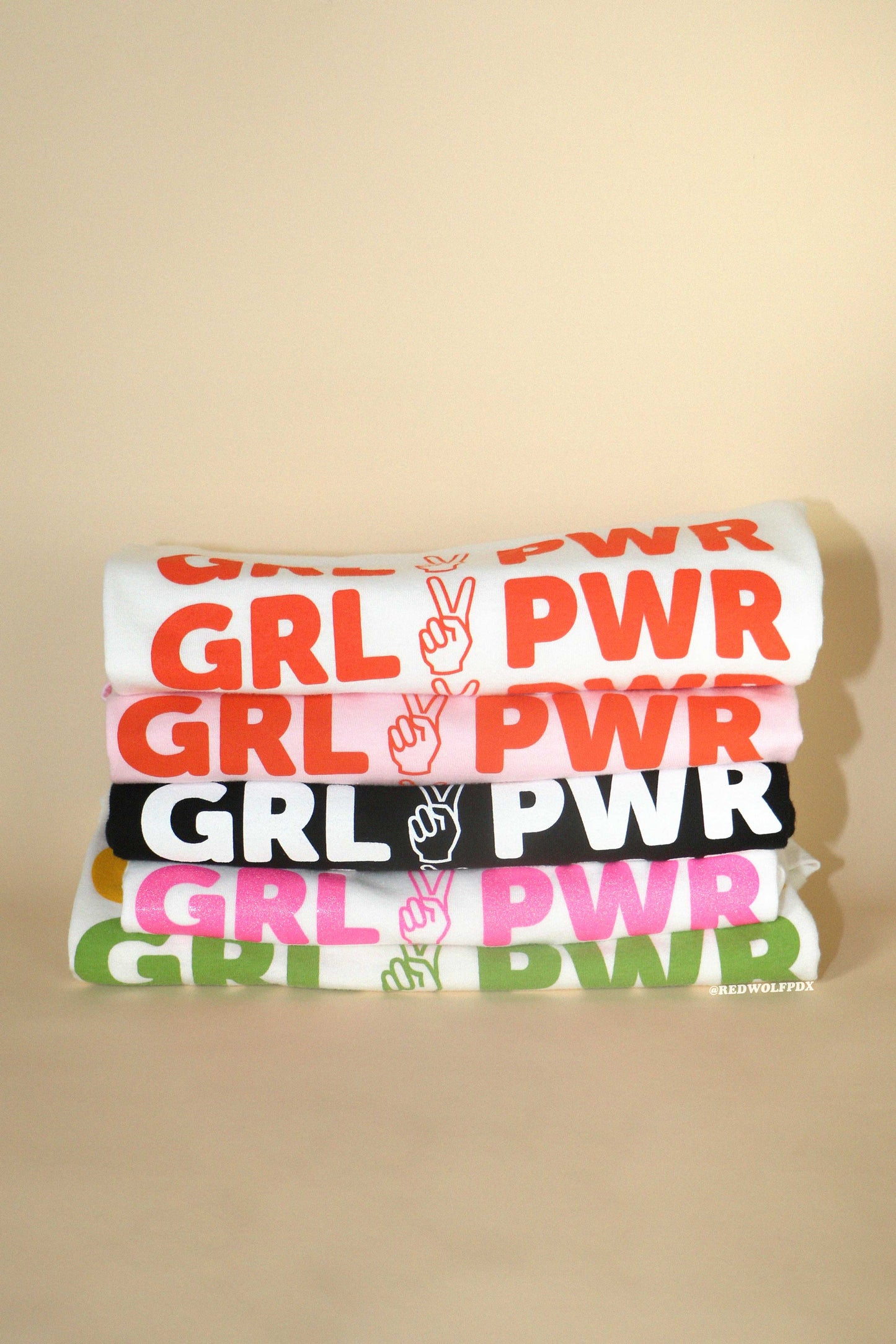 Stack of tees with 'GRL PWR' peace sign graphic screen printed across them on a beige background