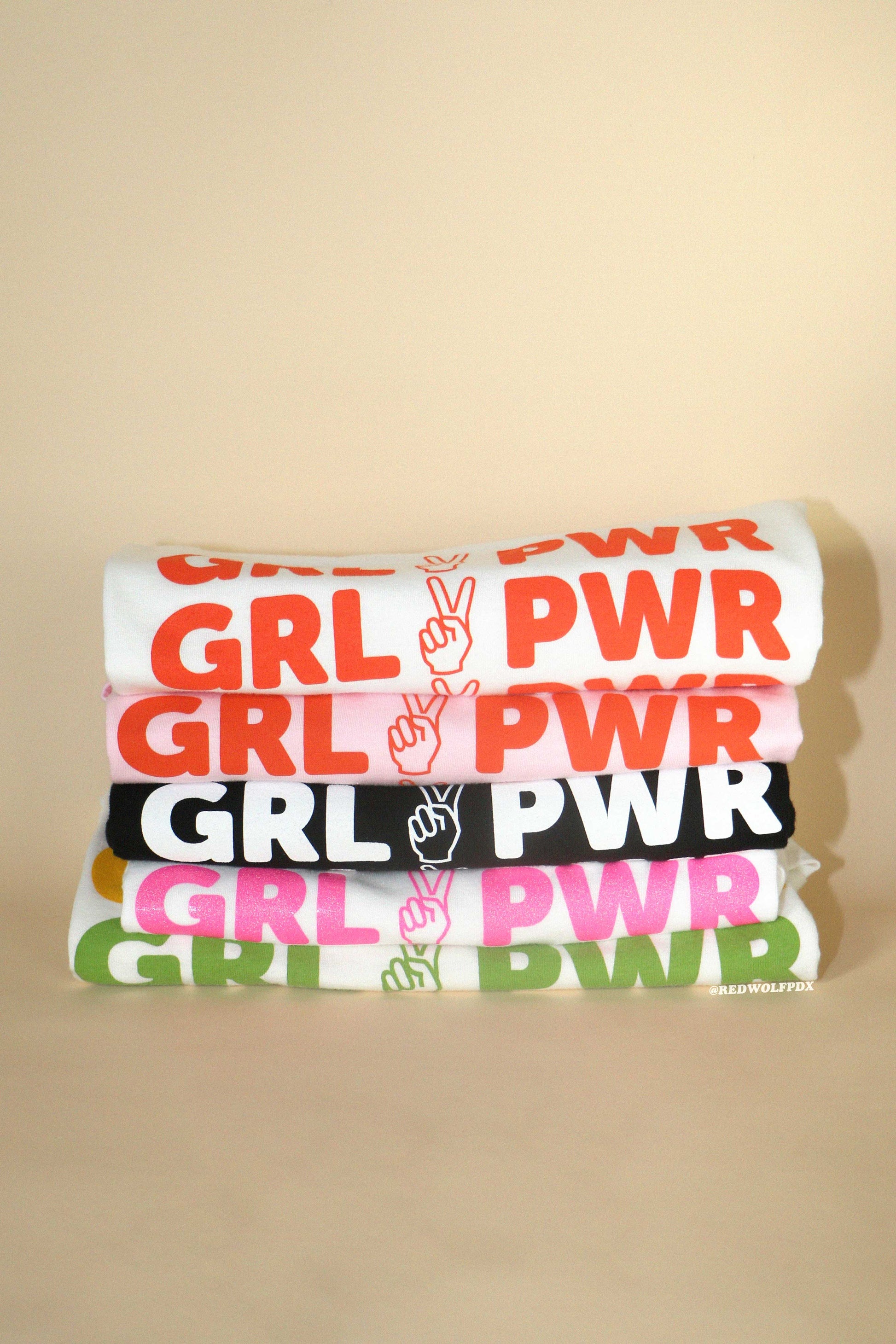 Stack of tees with 'GRL PWR' peace sign graphic screen printed across them on a beige background