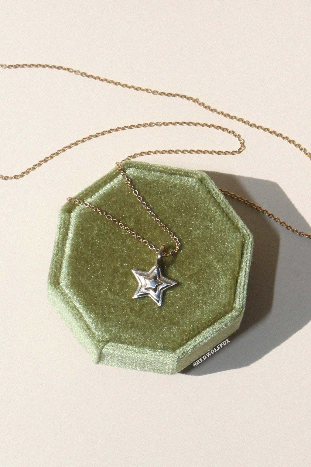 A brass starburst 3-d layered charm hanging from a brass chain necklace displayed on a green octagonal box on a beige background.